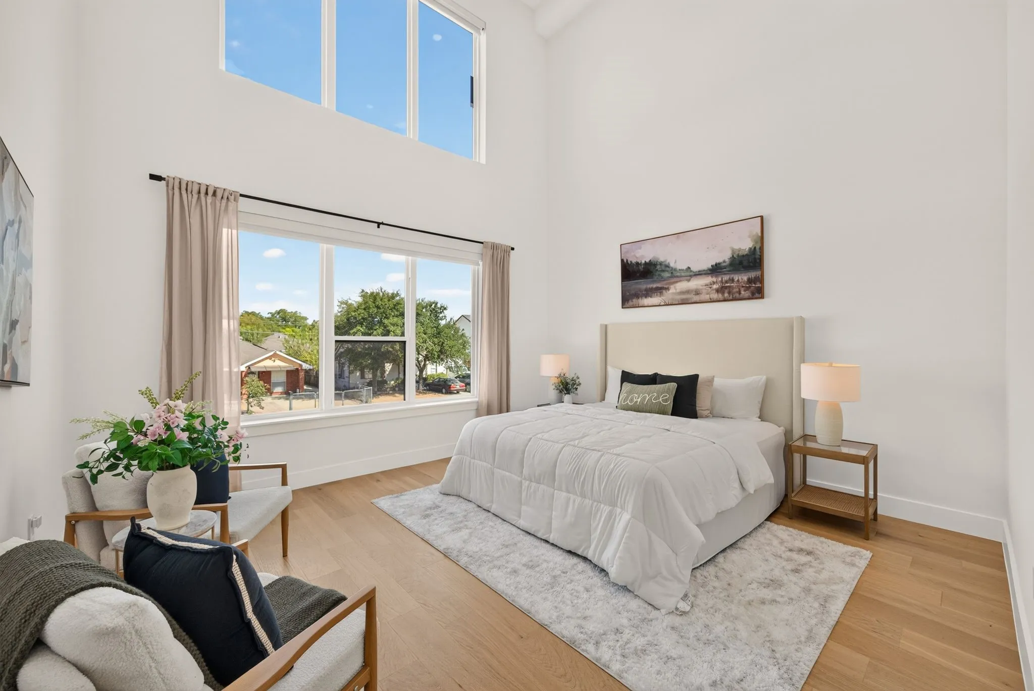 High ceilings with clerestory windows, expansive window overlooking neighborhood trees, wide-plank wood-finish flooring, white walls, and a light-colored upholstered headboard