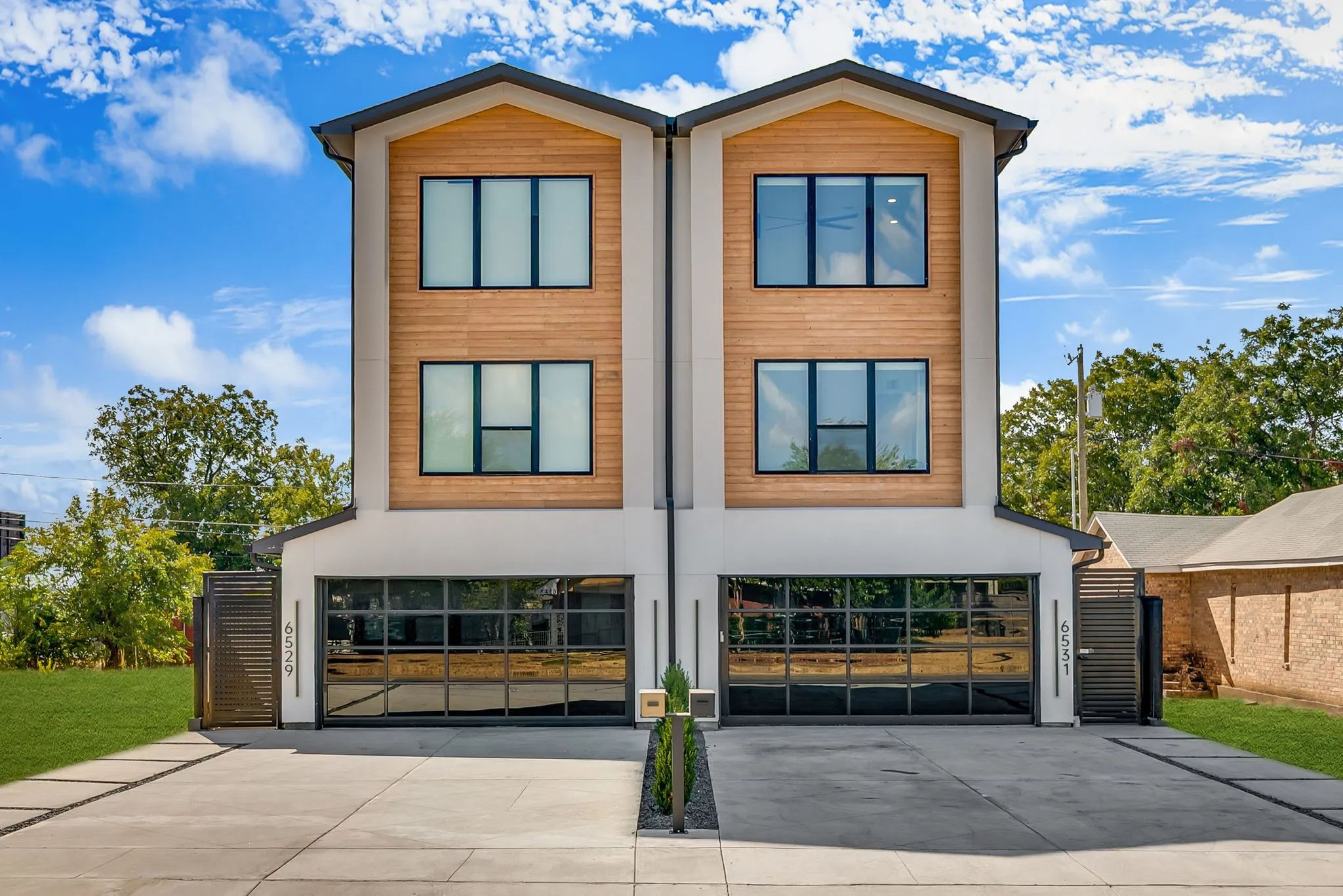 Modern duplex exterior featuring a clean white facade, natural wood-finish siding, black-framed windows, and glass garage doors