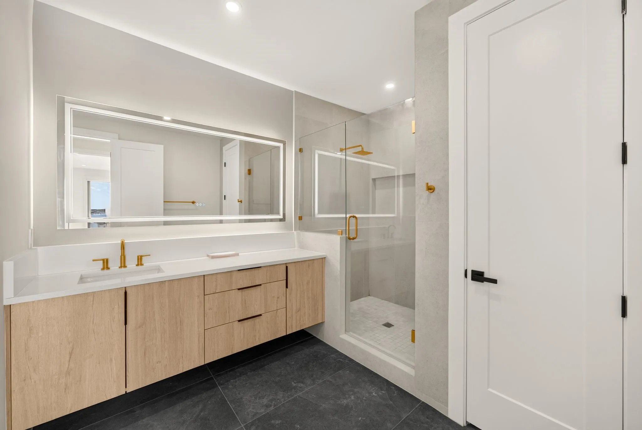 Floating wood-finish vanity with an integrated sink, illuminated mirror, and a glass-enclosed shower with gold-tone fixtures