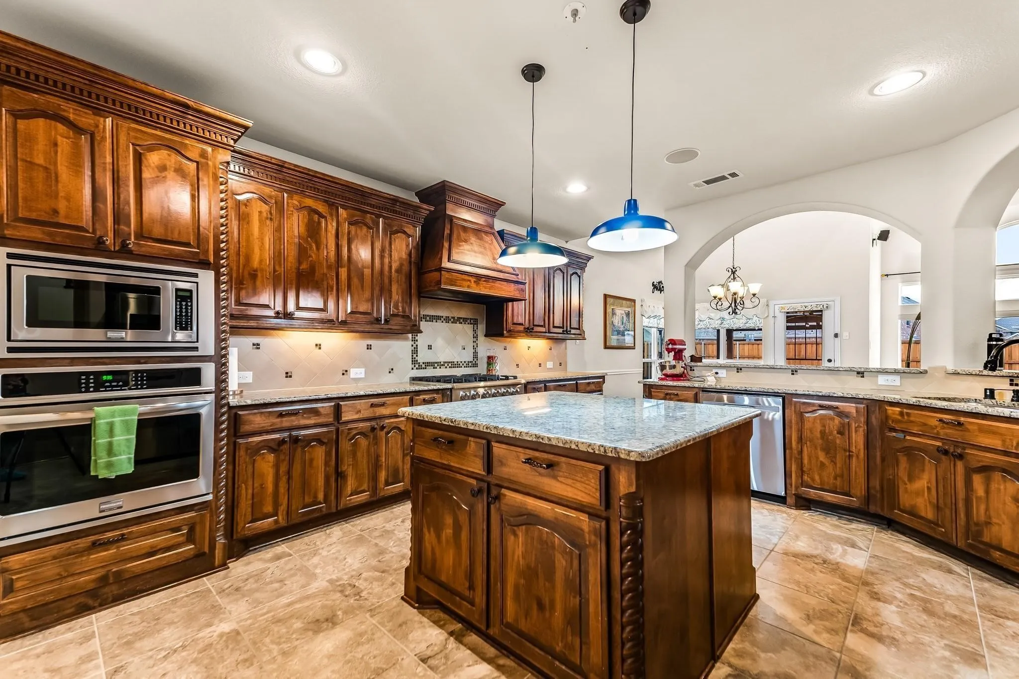Custom wood cabinetry with ornate detailing, stainless steel built-in appliances, recessed lighting, a central island with a light-toned countertop, and tile flooring
