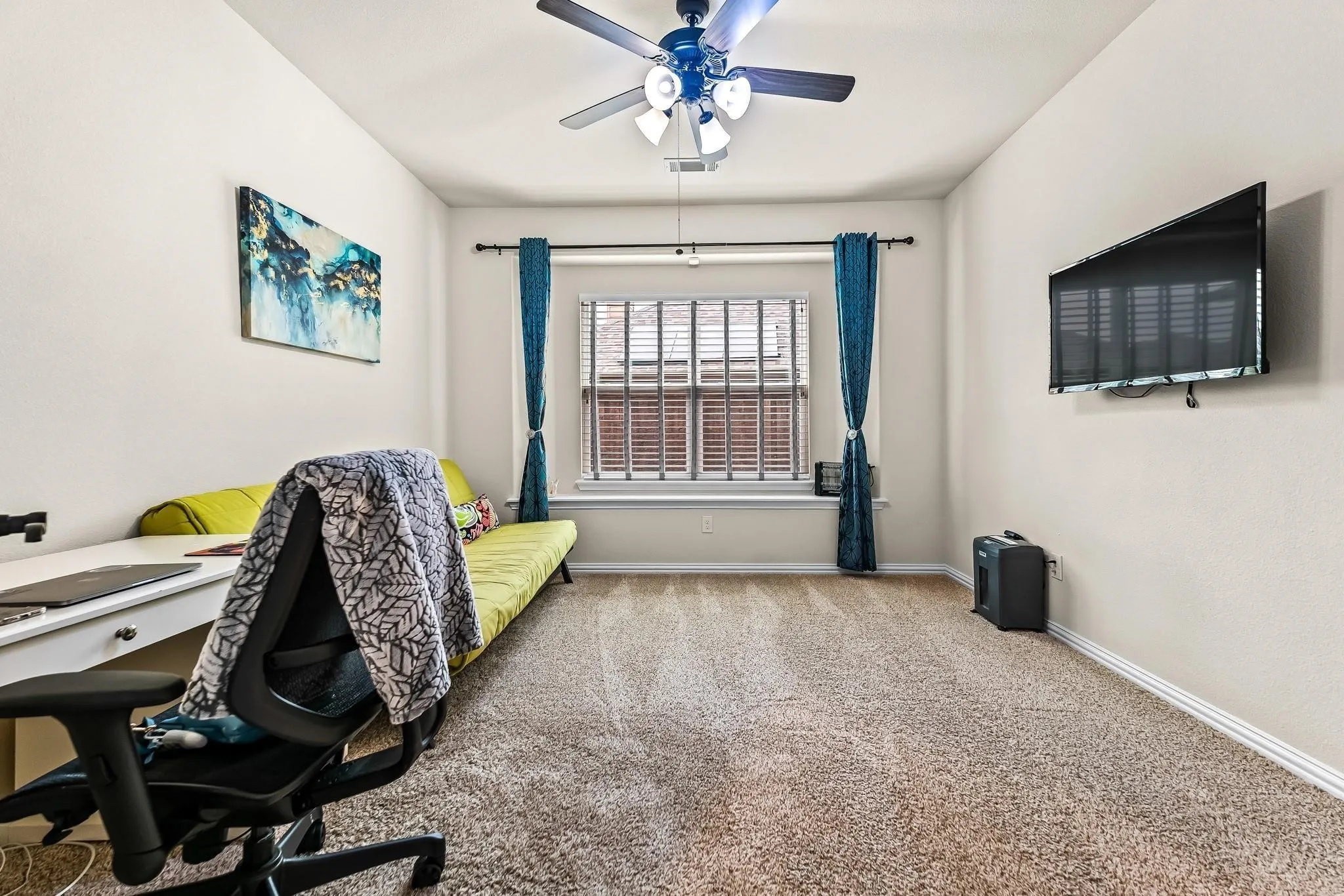 Window with blinds and security bars, wall-mounted television, ceiling fan with light fixture, light-colored carpeting, and neutral wall paint