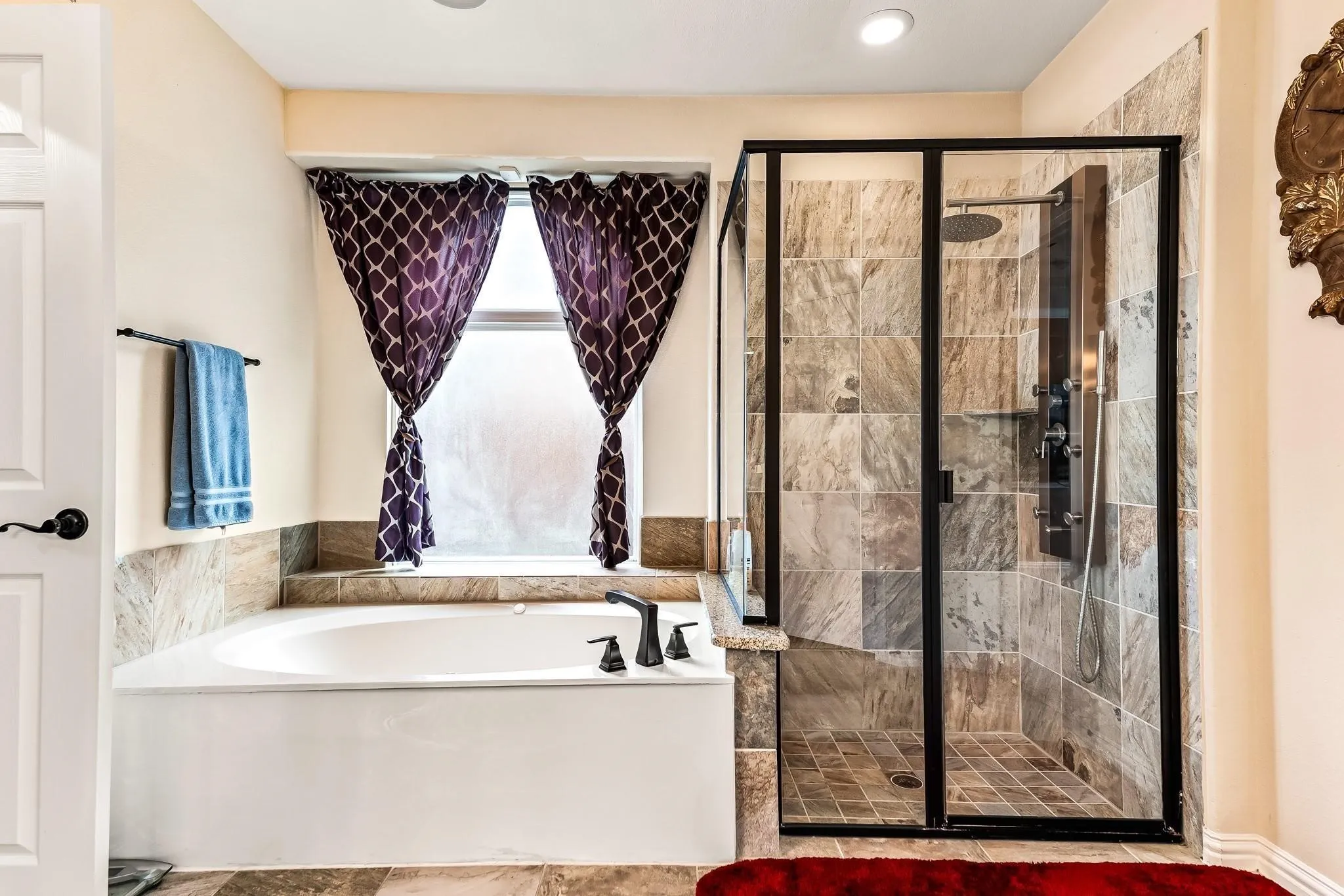 Built-in soaking tub with dark bronze fixtures, frosted window, and adjacent tiled shower with a multi-function shower panel