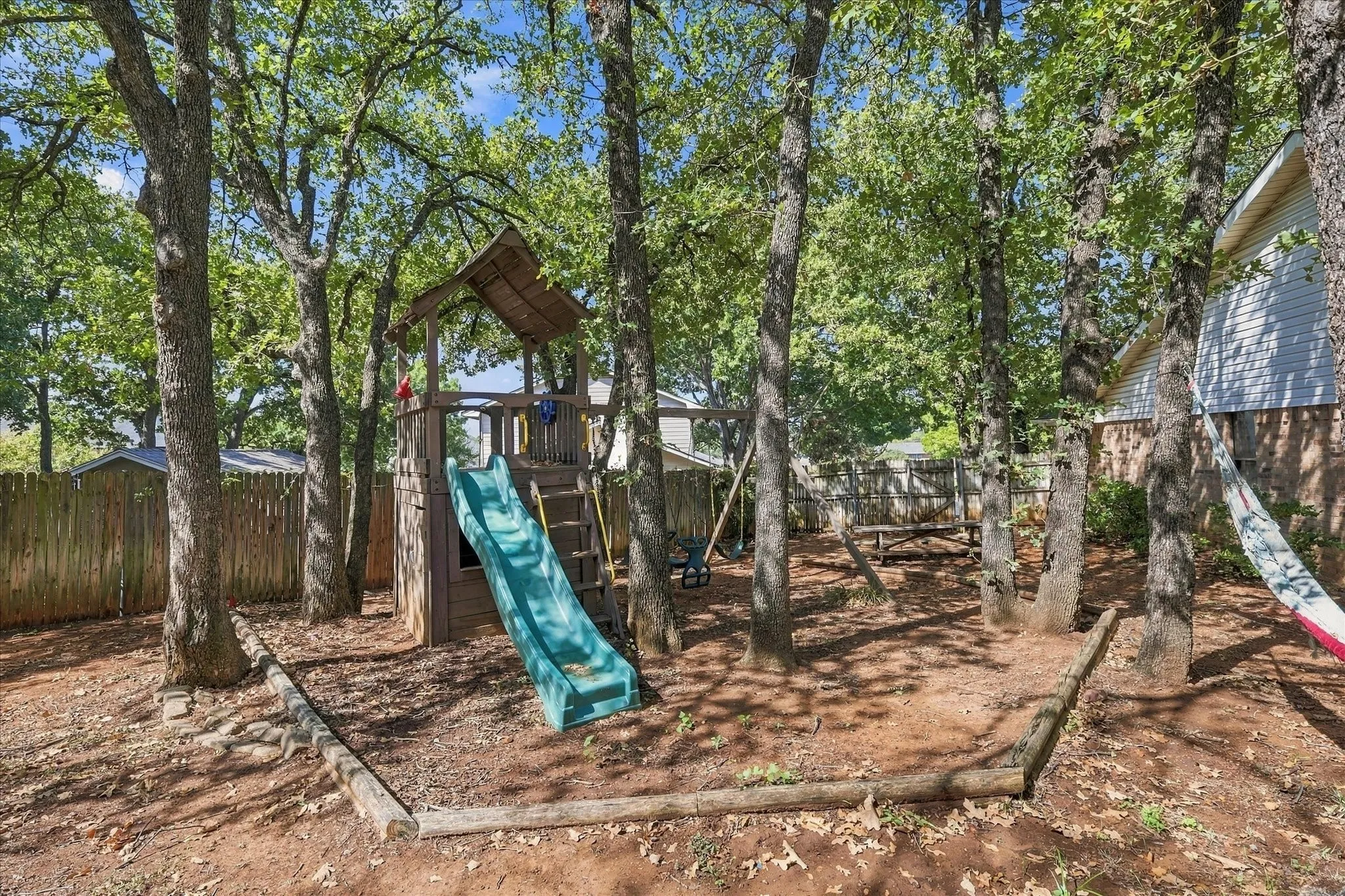 Fenced backyard featuring mature trees, a built-in wooden play structure with slide, and a swing set