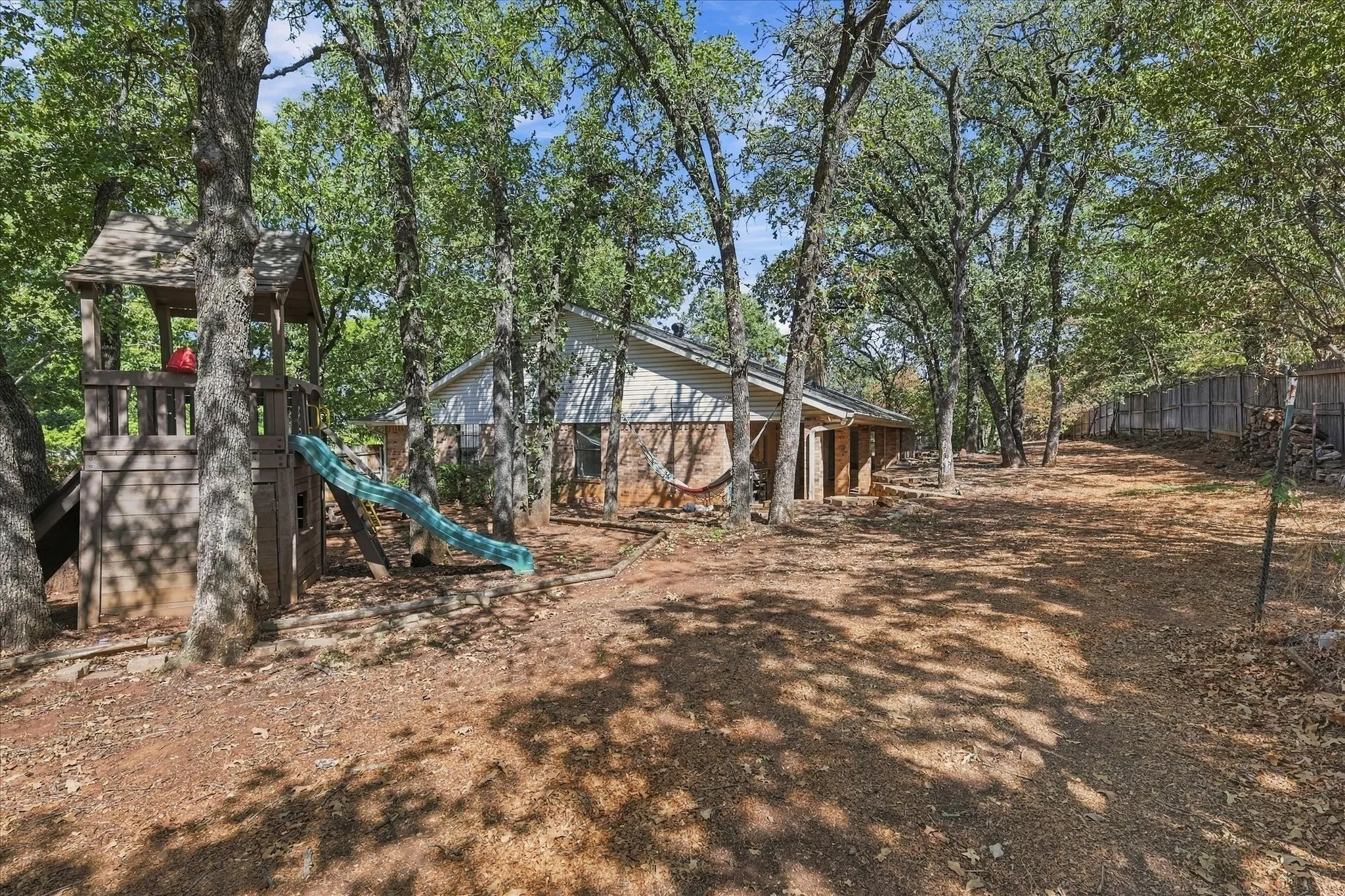 Expansive backyard featuring mature trees, a wooden play structure with a slide, a partial privacy fence, and a brick exterior on the residence