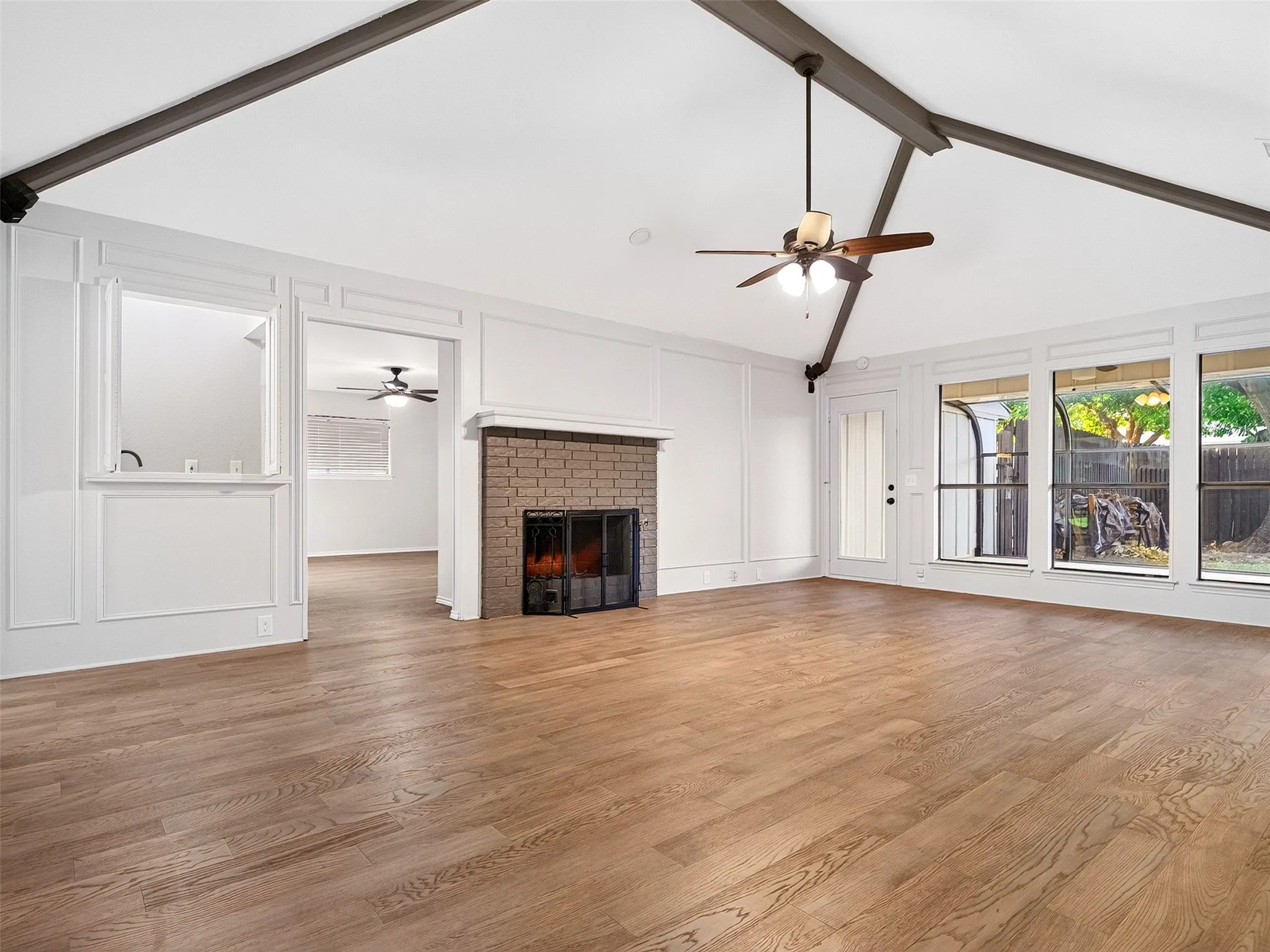 Vaulted ceiling with exposed beams, wood-finish flooring, brick-faced fireplace with hearth, ceiling fan light fixture, and large windows