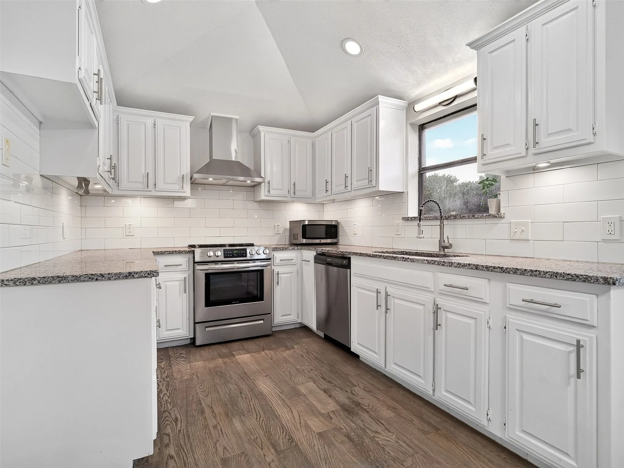White cabinetry with brushed nickel hardware, stainless steel appliances, granite countertops, white subway tile backsplash, and wood-finish flooring