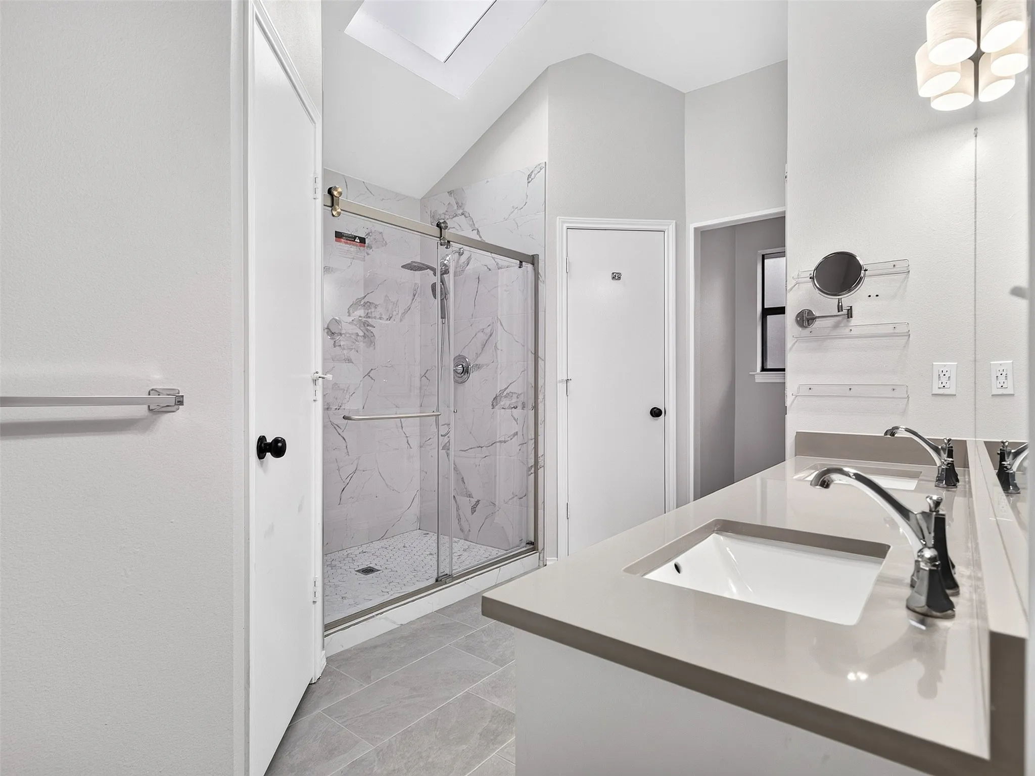 Dual vanity with integrated sinks, a glass-enclosed shower with tile surround, a skylight, and a contemporary ceiling fixture