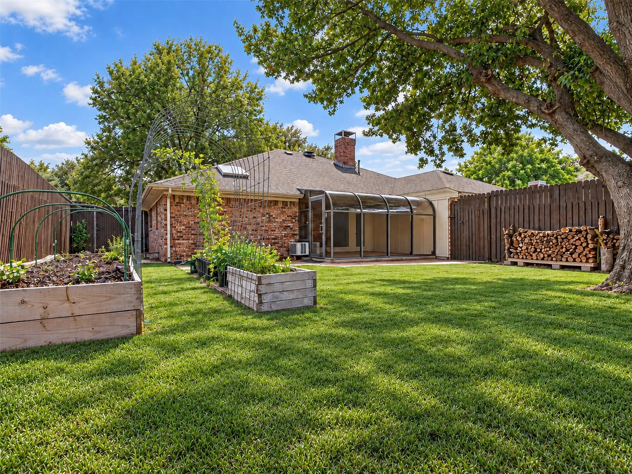 Expansive backyard featuring a manicured lawn, brick exterior, enclosed patio with a translucent roof, a wood privacy fence, and raised garden beds