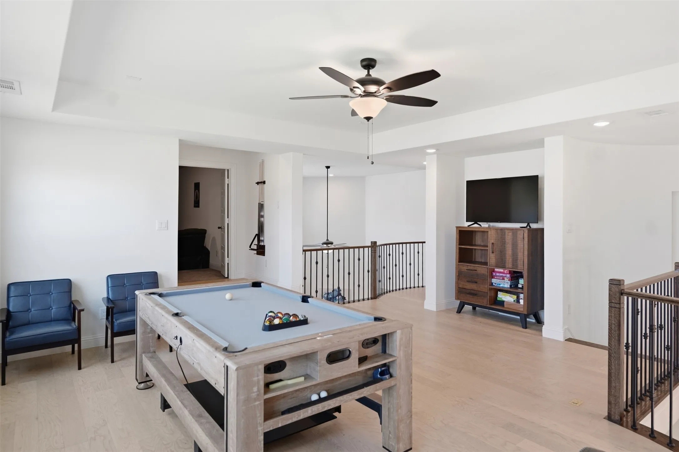 Open loft area with wood-finish flooring, ceiling fan with integrated lighting, and wrought iron balusters