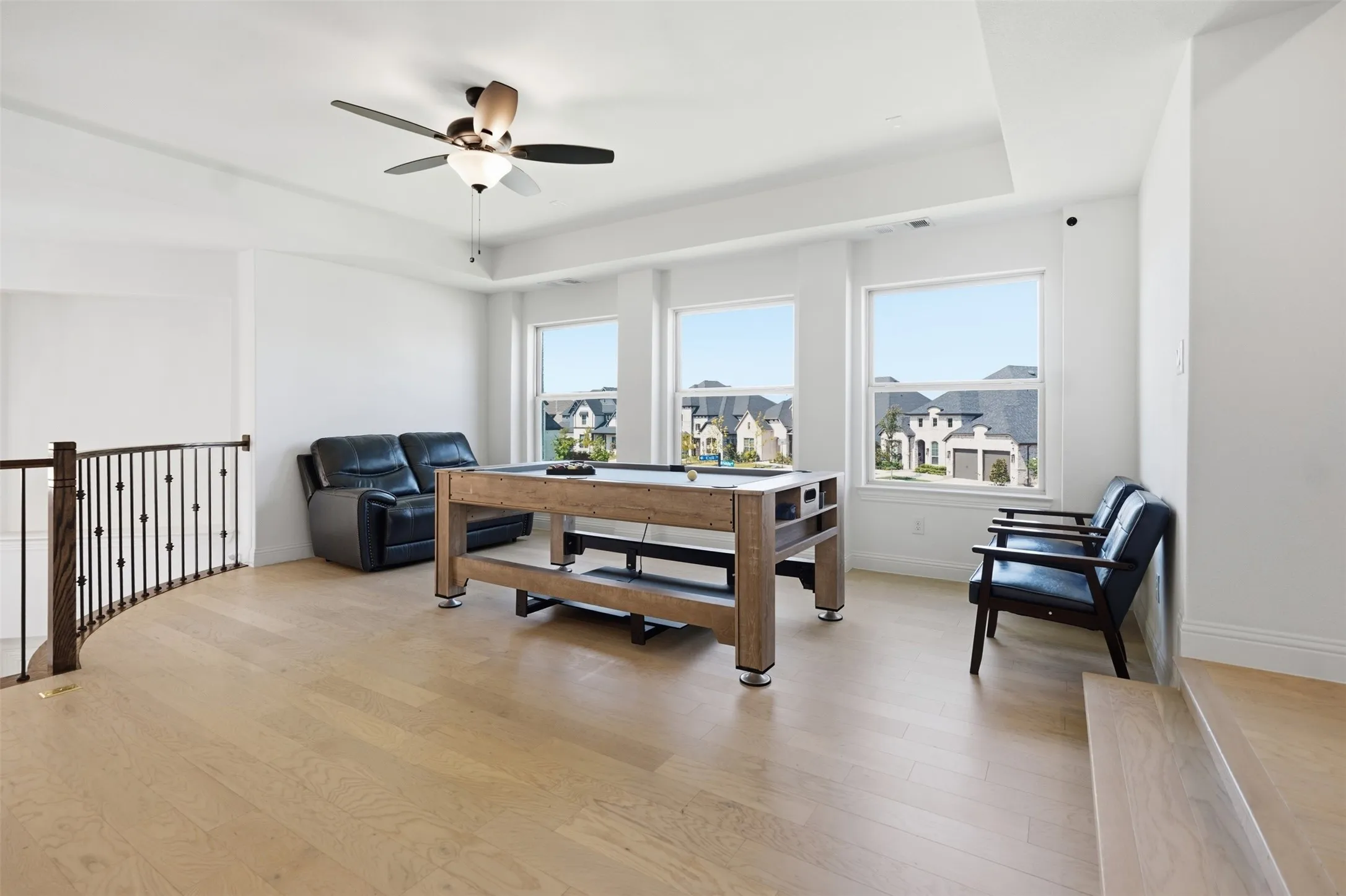 Spacious loft area featuring light wood-finish flooring, three windows, a tray ceiling with a ceiling fan, and wrought iron balusters with a wood handrail