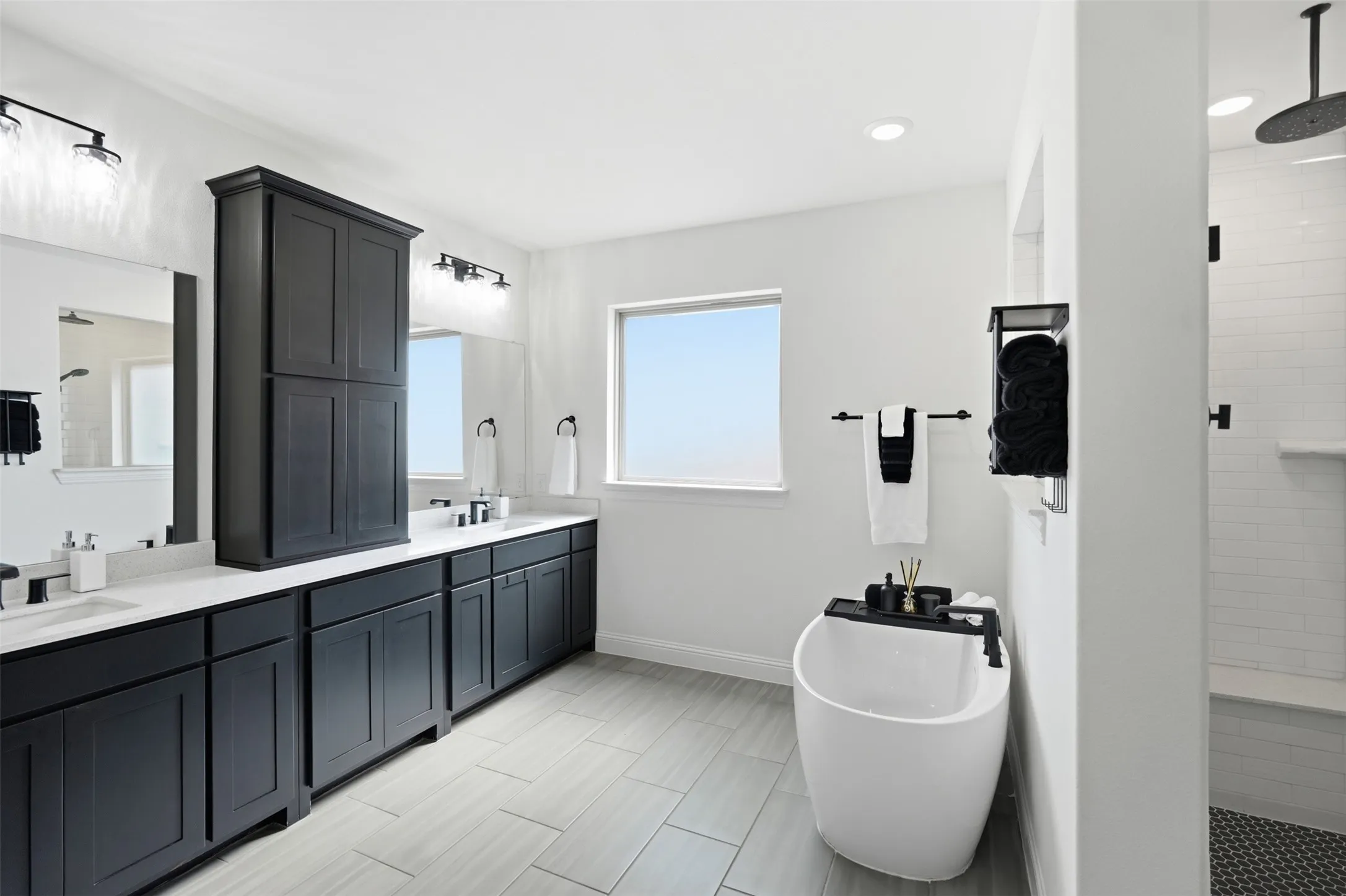 Double vanity with dark cabinetry and white countertops, freestanding soaking tub, separate shower with tiled walls, and light-toned tile flooring