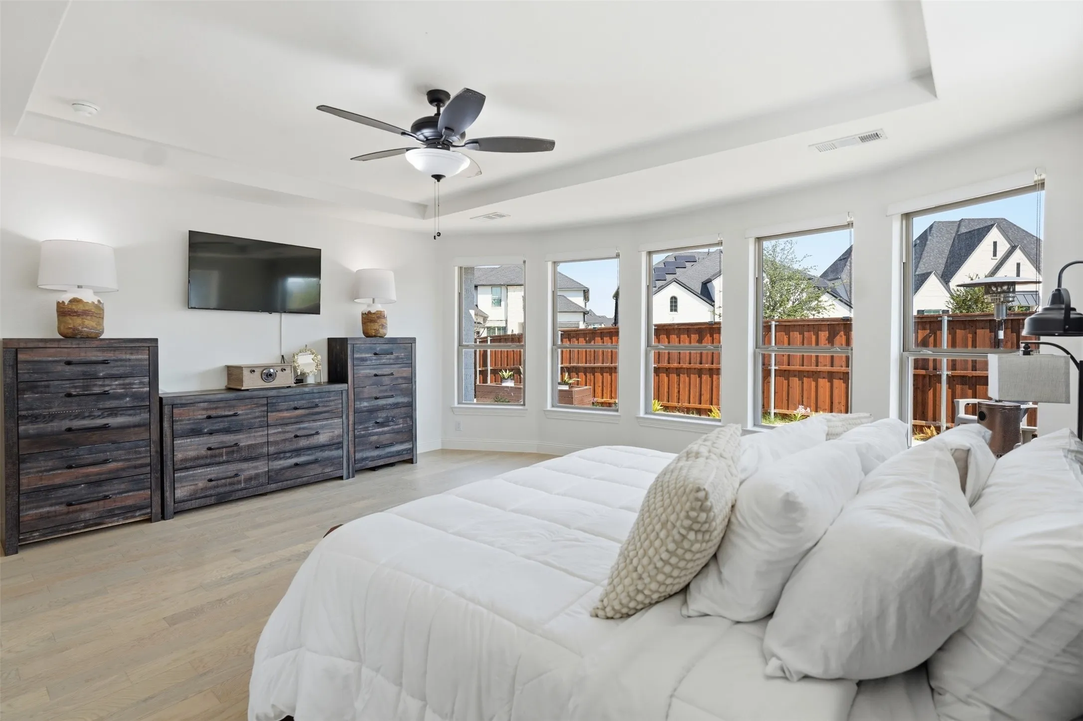 Tray ceiling with a ceiling fan, light wood-finish flooring, and multiple windows with a privacy fence