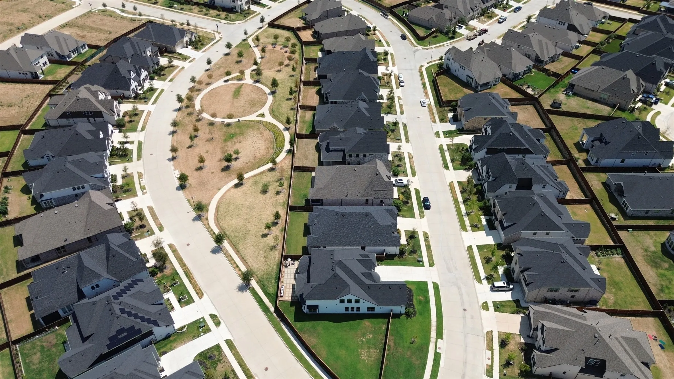 Residential development featuring individual homes with dark-toned roofs, concrete streets, and a central landscaped green space with walking paths