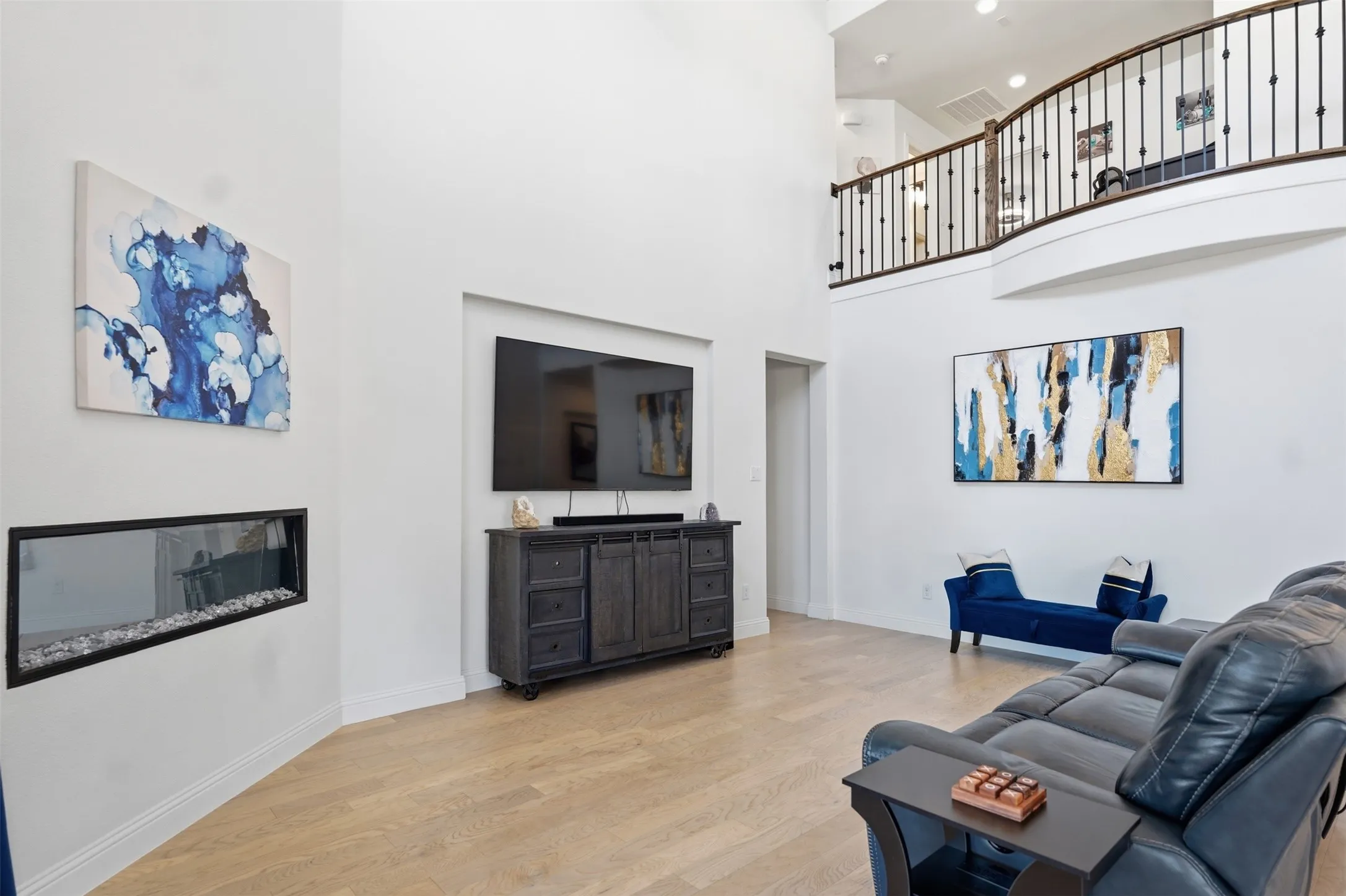 Two-story great room featuring a linear built-in fireplace, wood-finish flooring, and a second-story balcony with wrought iron railing