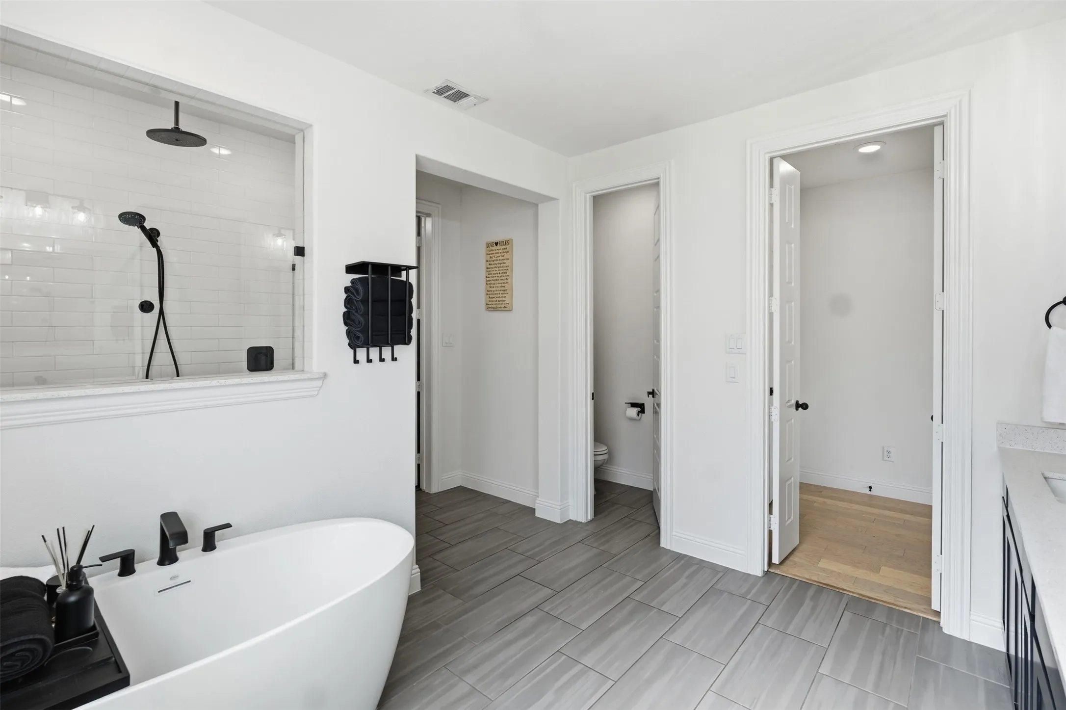 Freestanding soaking tub with matte black fixtures, glass-enclosed shower with subway tile surround, matte black rain shower head and handheld sprayer, wood-finish flooring, and light gray plank tile flooring