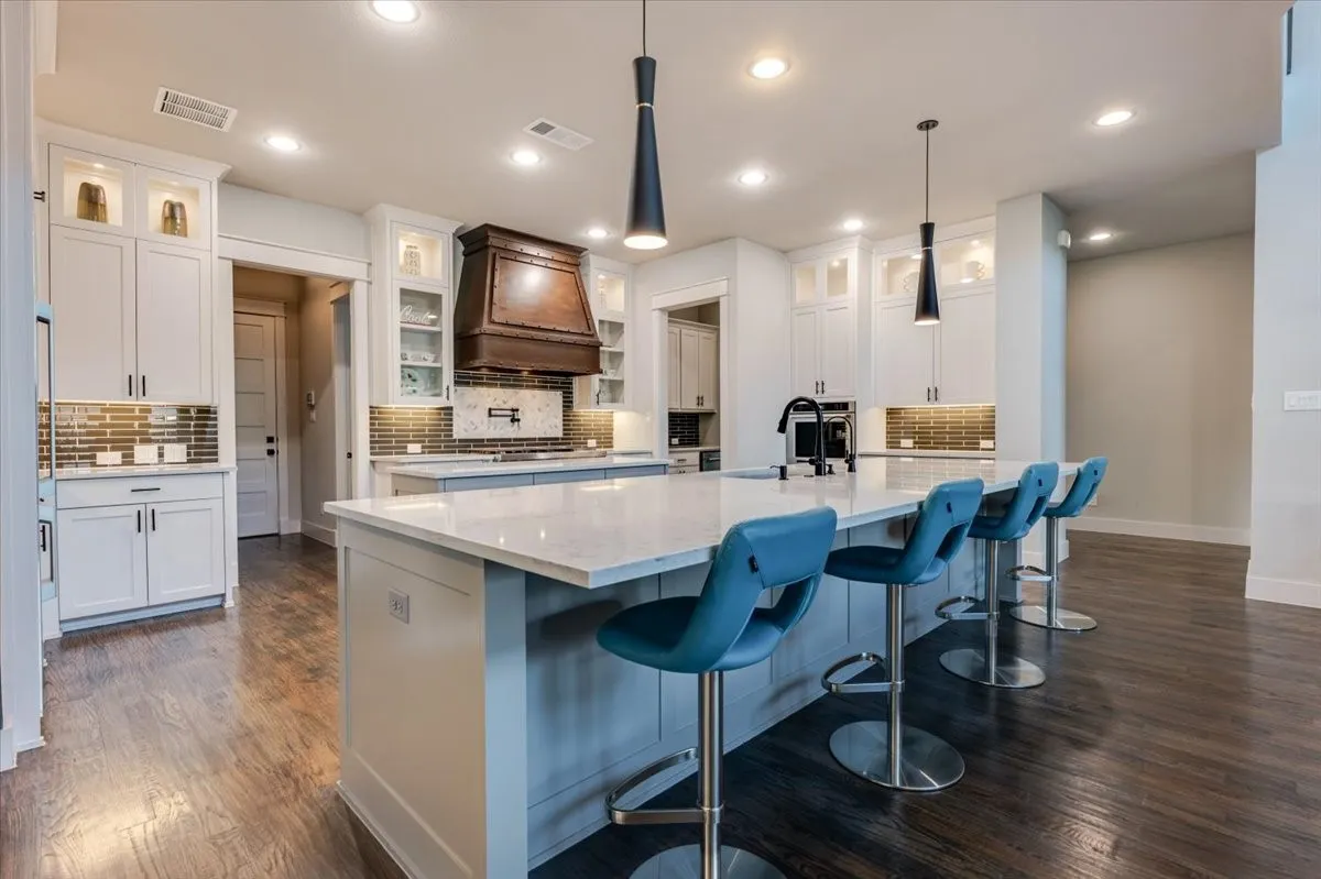 Spacious kitchen featuring an expansive central island with a light-toned countertop, white cabinetry with display lighting, a tiled backsplash, and wood-finish flooring