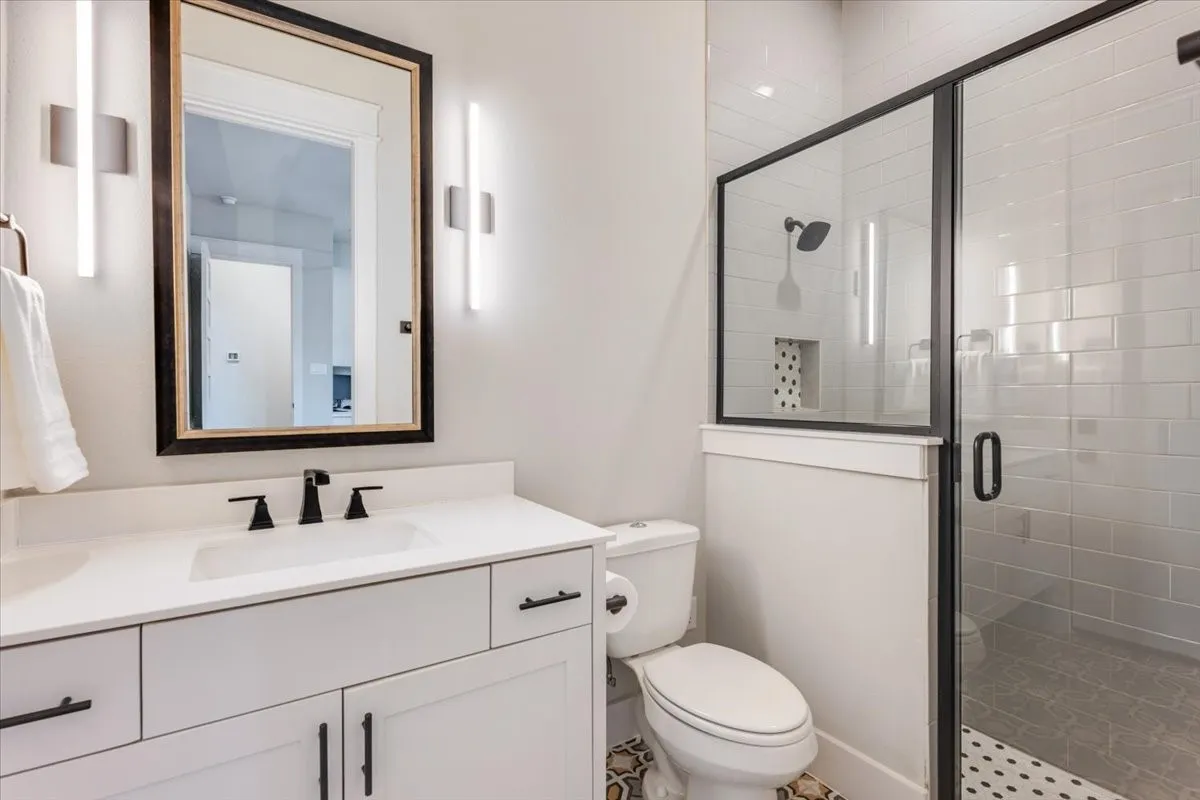 Modern bathroom featuring a white vanity with dual drawers and matte black hardware, white quartz countertop, framed mirror with integrated lighting, and a walk-in shower with subway tile surround and frameless glass enclosure