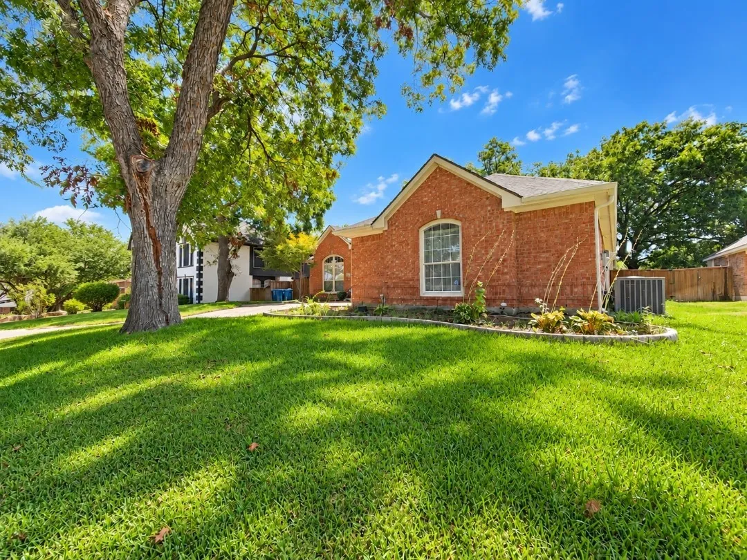 Brick exterior with arched window, established front tree, landscaped garden bed, and a verdant lawn