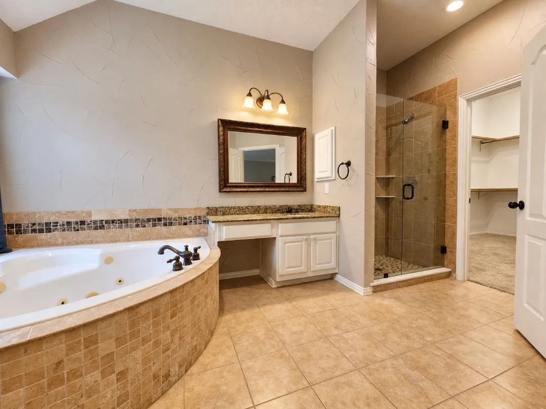 Deep soaking tub with tiled surround, glass-enclosed shower with tiled walls and pebble floor, vanity with granite countertop and integrated sink, recessed lighting, and tile flooring