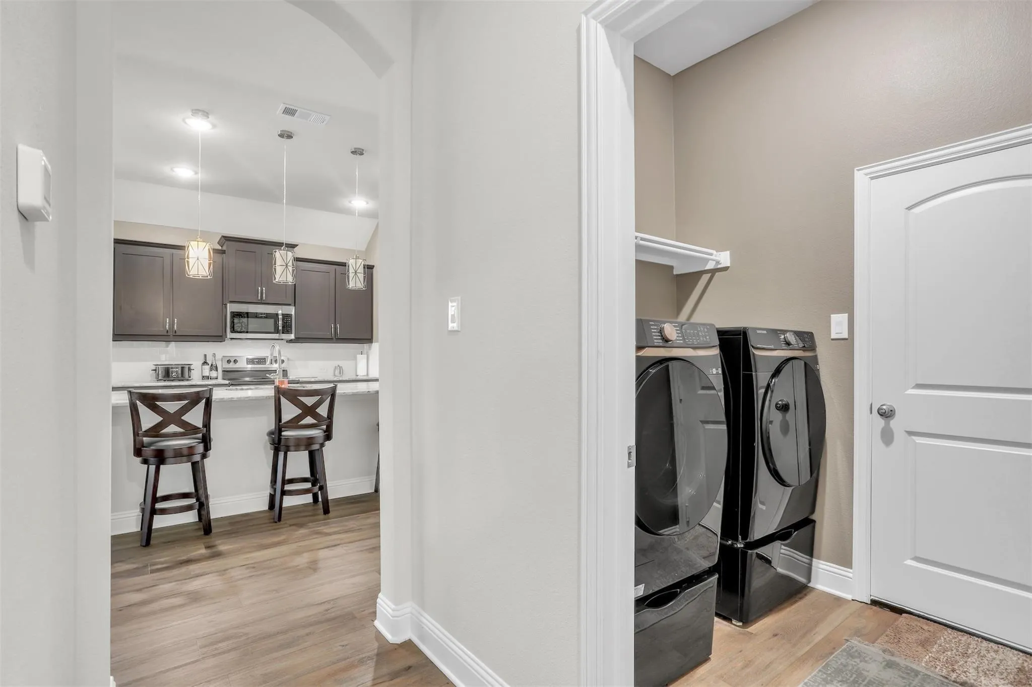 Wood-finish flooring throughout, kitchen featuring dark wood cabinetry and pendant lighting, white countertop, stainless steel microwave, and laundry space with shelf