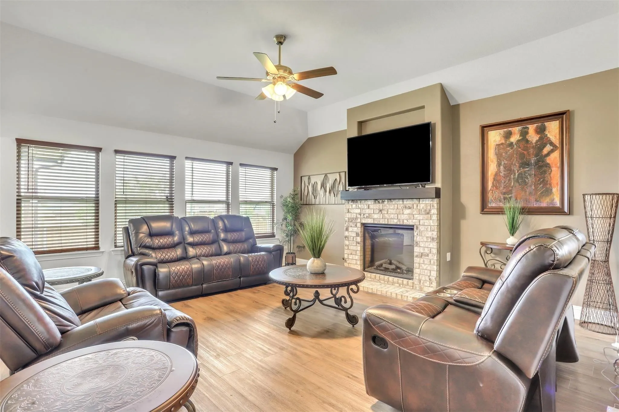 Vaulted ceiling, wood-finish flooring, multiple windows with blinds, brick-surround fireplace, and ceiling fan with integrated lighting