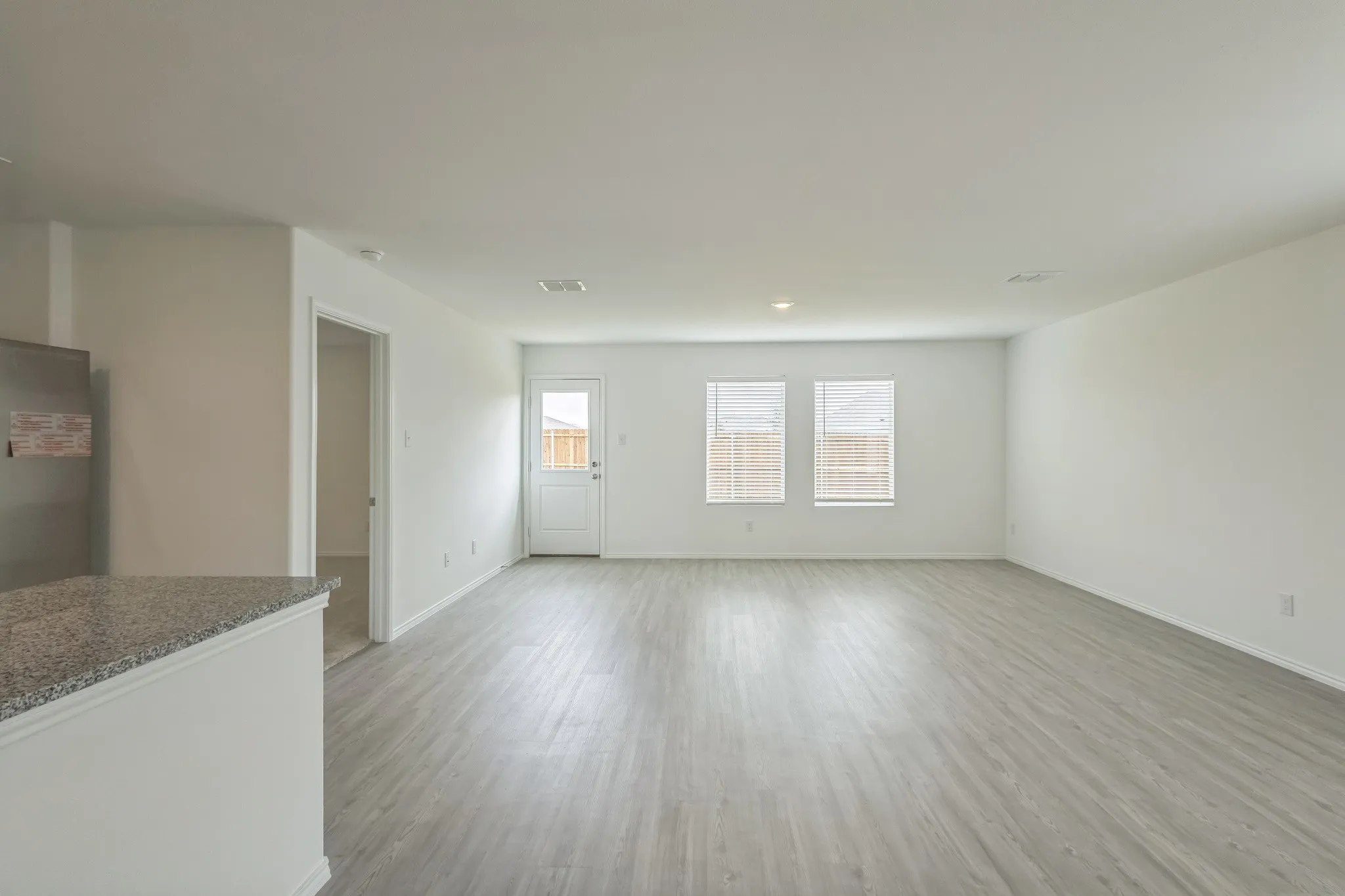 Spacious open-concept area featuring wood-finish flooring, white walls, multiple windows with blinds, and a rear exterior door