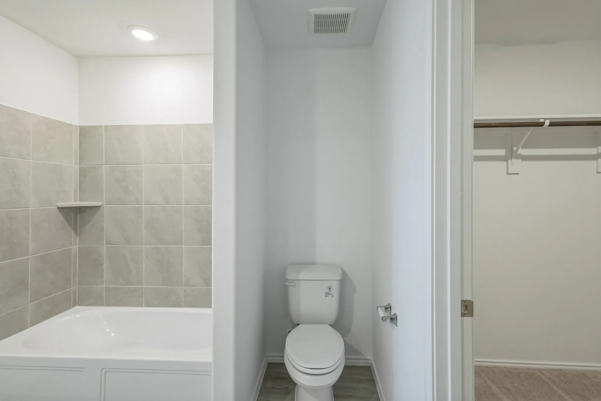 Bathtub with tiled surround and integrated shelf, recessed lighting, toilet with wall-mounted toilet paper holder, wood-finish flooring, and a closet with a hanging rod