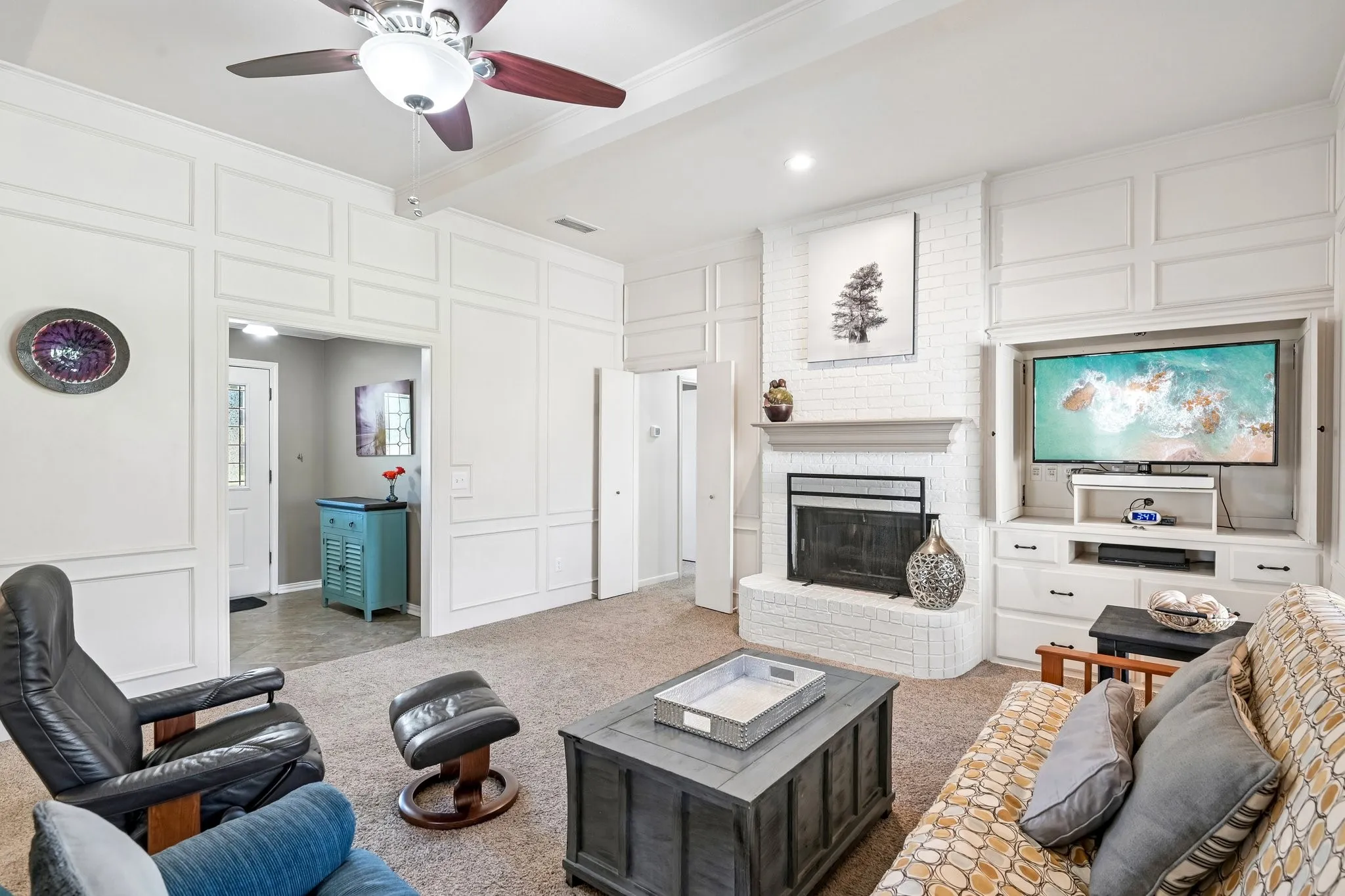 White paneled walls, ceiling fan with light fixture, brick fireplace with mantel, built-in entertainment center, and carpeted flooring