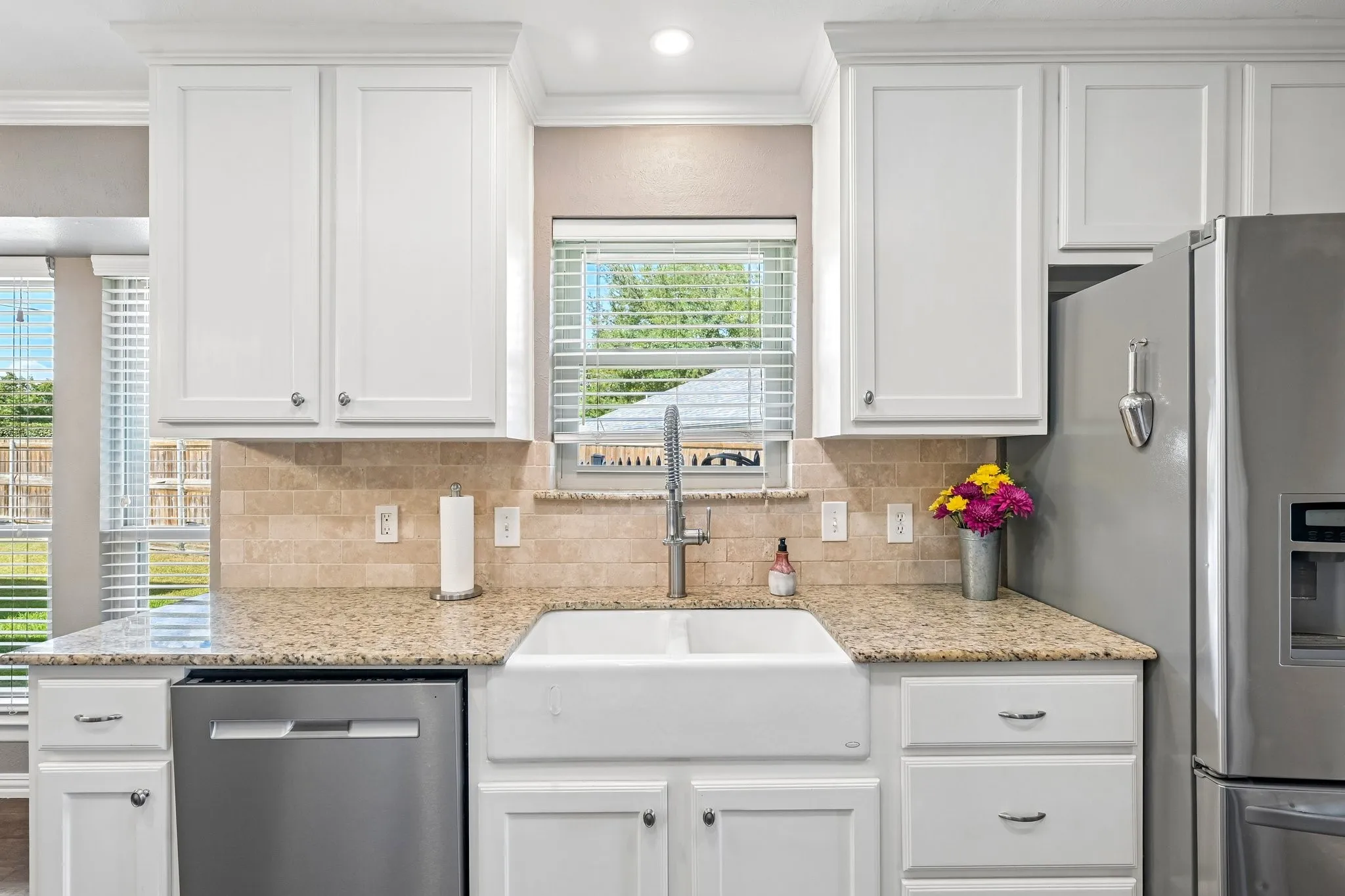 White shaker-style cabinetry with crown molding, light-toned granite countertops, a farmhouse-style sink, and a tiled backsplash