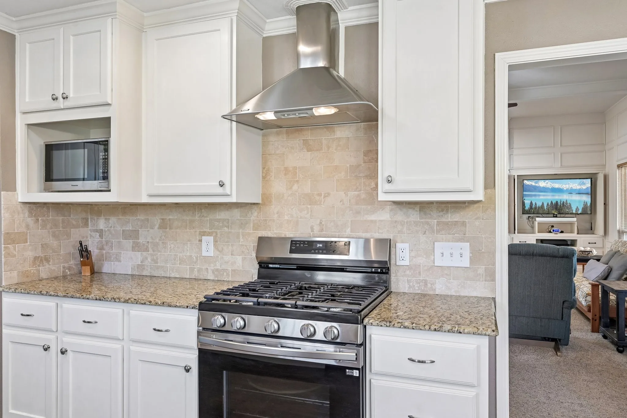 White cabinetry with crown molding, stainless steel range with overhead hood, integrated microwave, granite-finish countertops, and natural stone tile backsplash