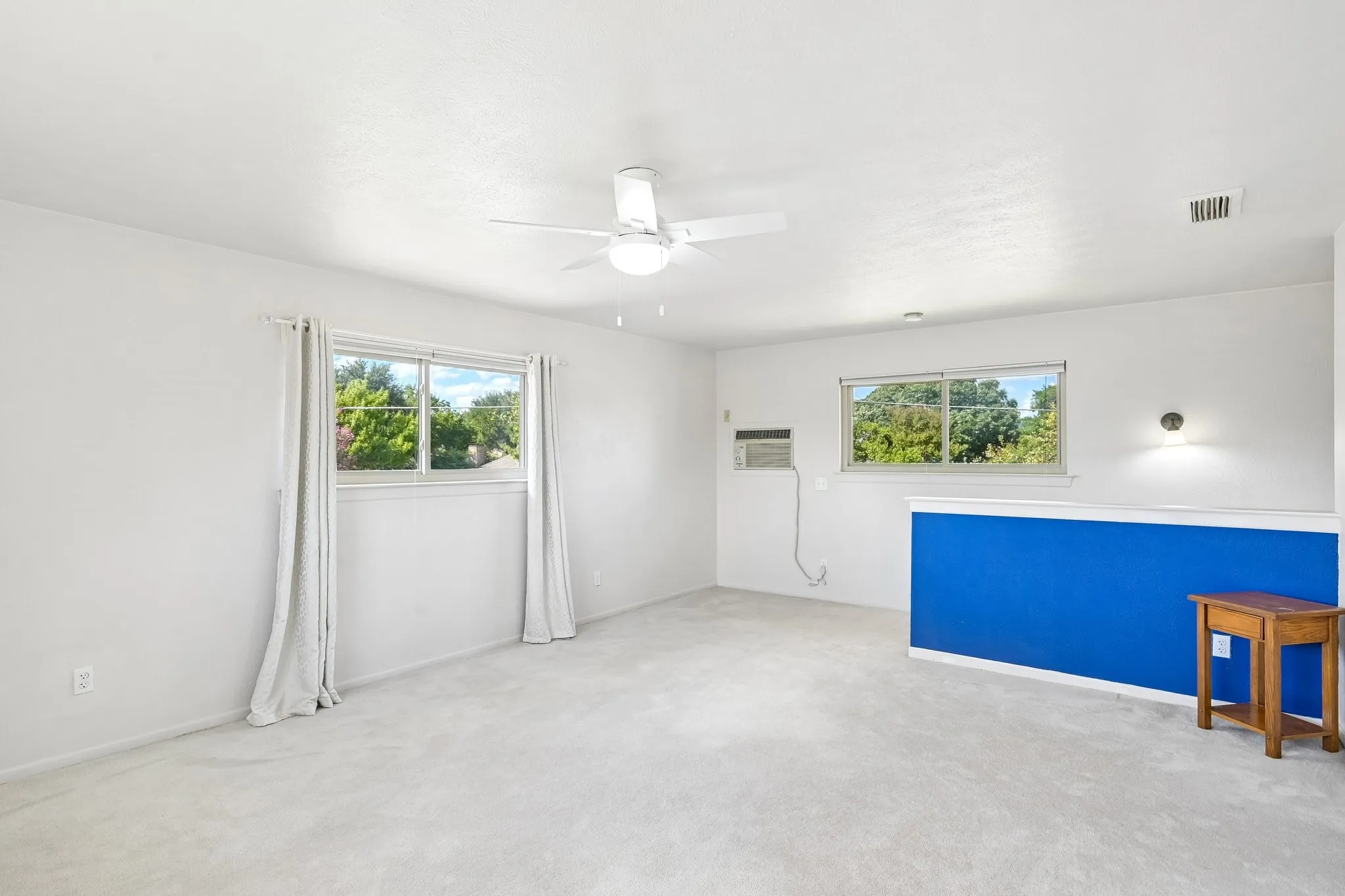 Expansive room featuring light-toned carpeting, multiple windows, a ceiling fan with integrated lighting, and a wall-mounted sconce