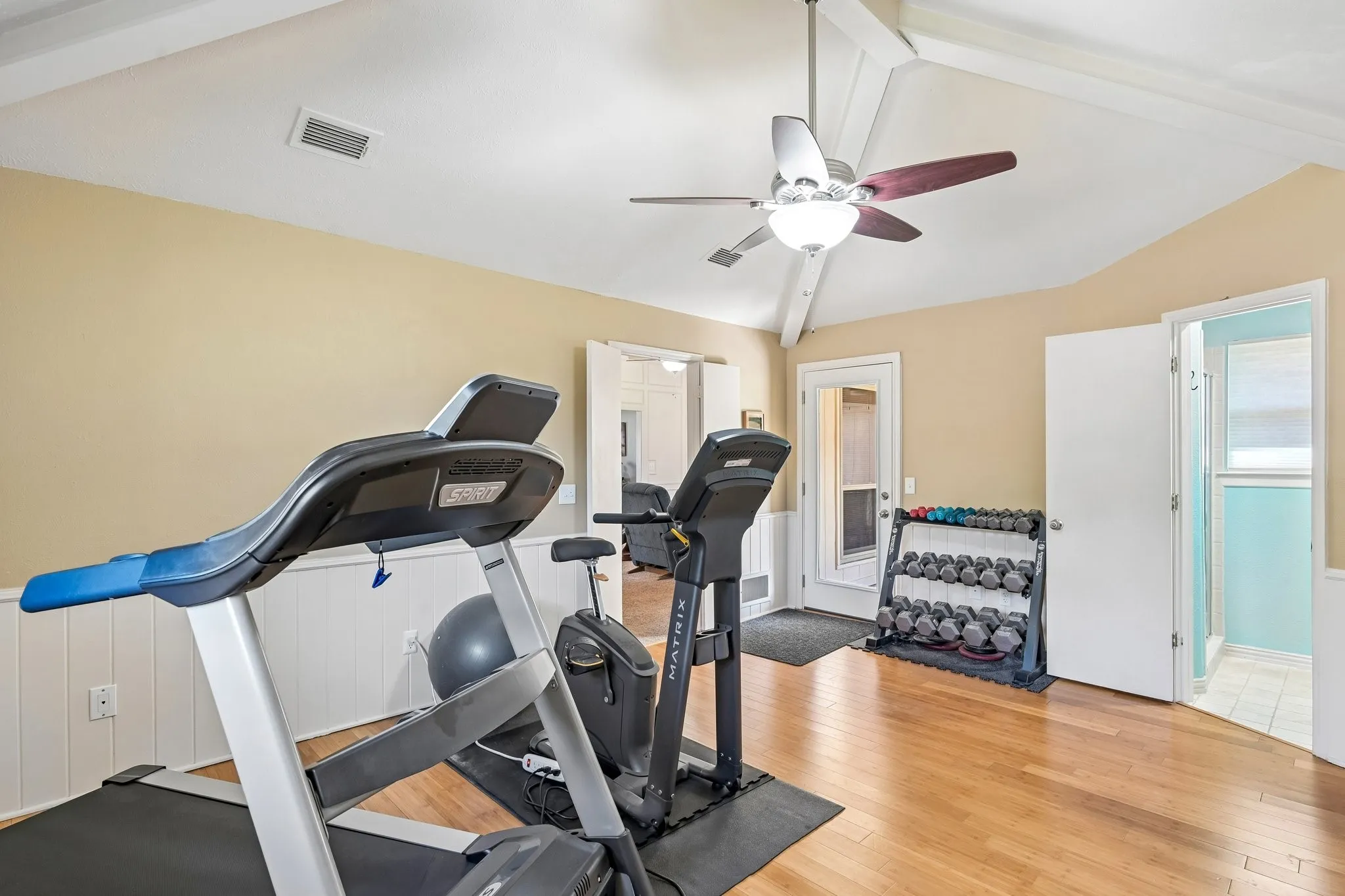 Vaulted ceiling with exposed beams, wood-finish flooring, beadboard wainscoting, and a ceiling fan with integrated lighting