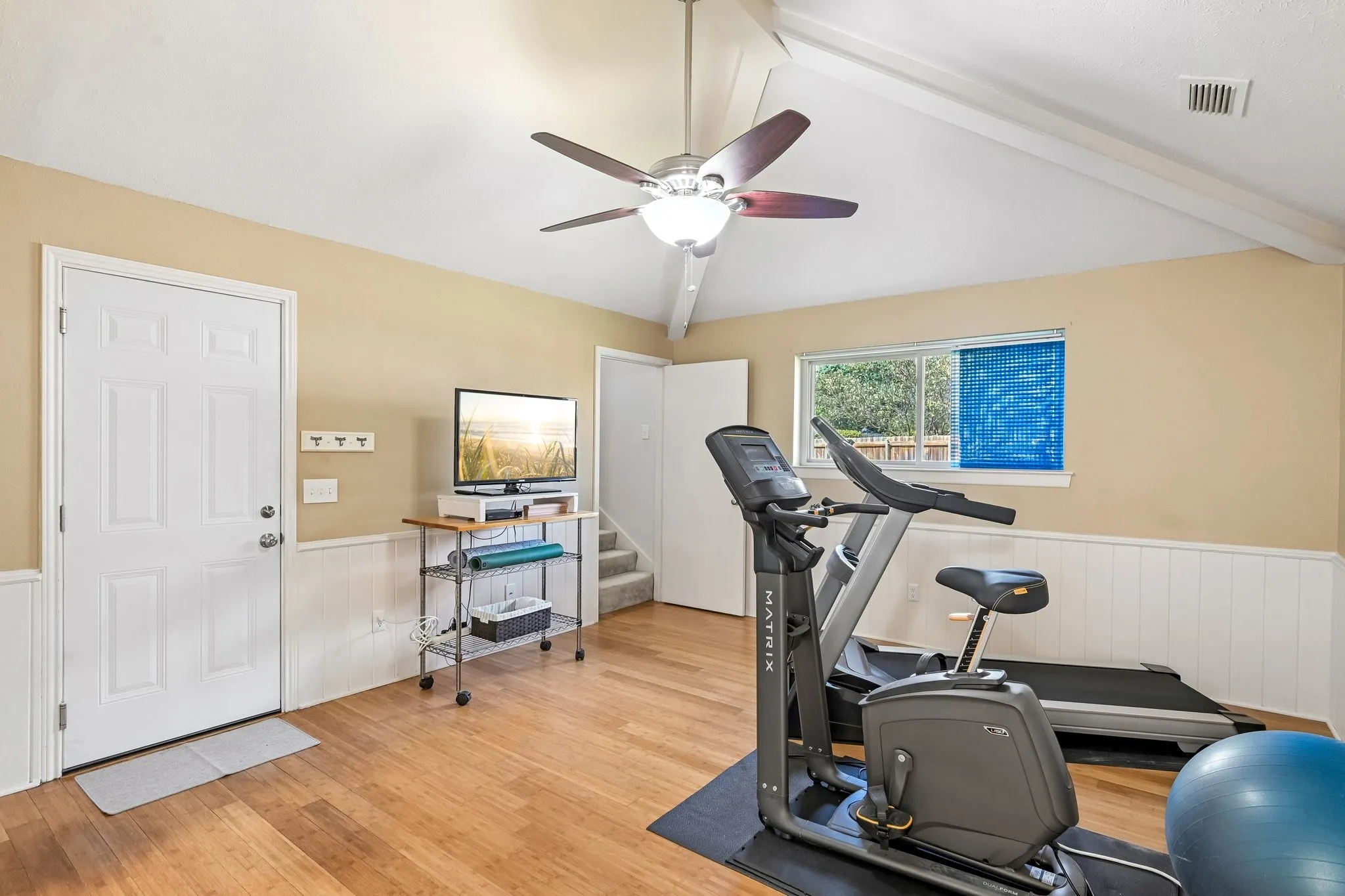 Vaulted ceiling with exposed beams, wood-finish flooring, half-wall wainscoting, and a ceiling fan