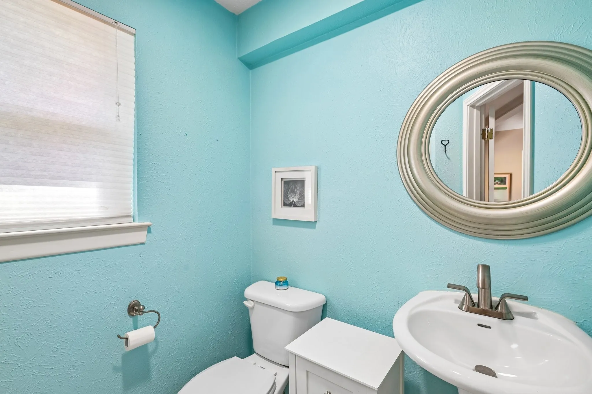 Pedestal sink with a brushed nickel faucet, coordinating oval mirror, and a window with pleated shades