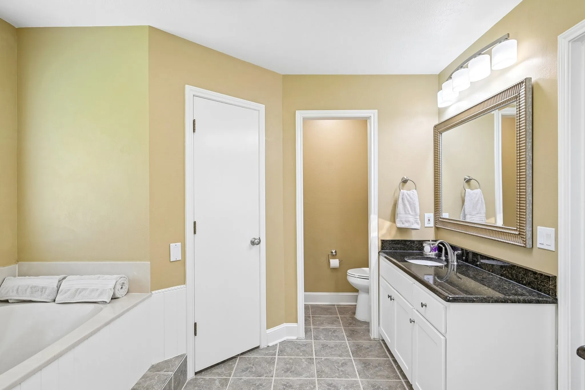 Spacious bathroom featuring a built-in bathtub, white vanity with ample storage, dark countertop, and a large framed mirror with overhead lighting