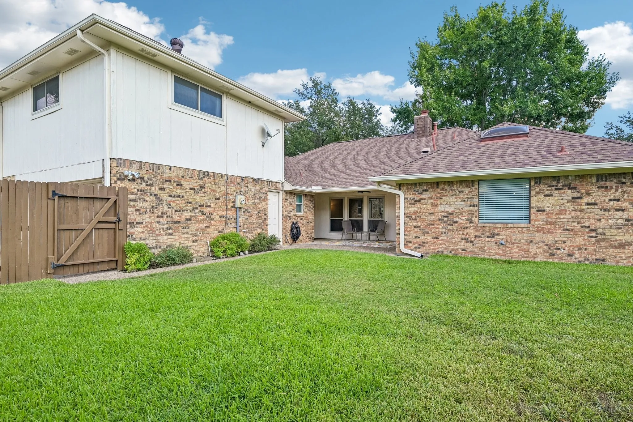 Expansive green lawn, multi-level brick and siding exterior, privacy fencing, covered patio, and a skylight feature