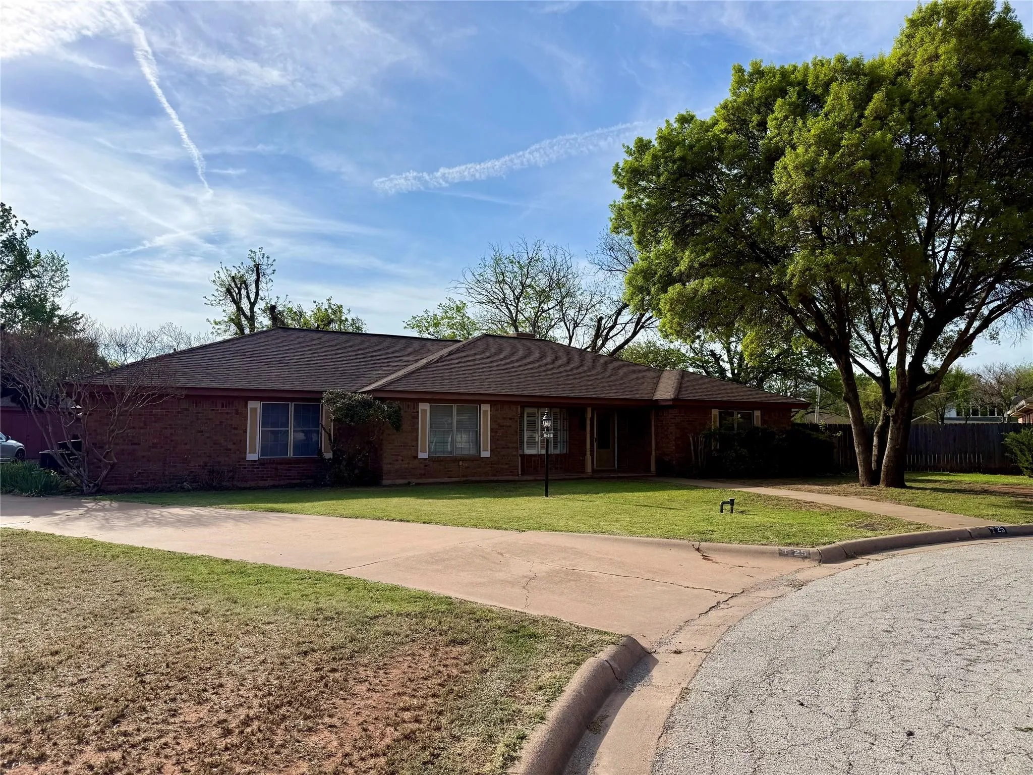 Brick exterior with a brown shingle roof, mature front yard tree, concrete driveway, and front walkway