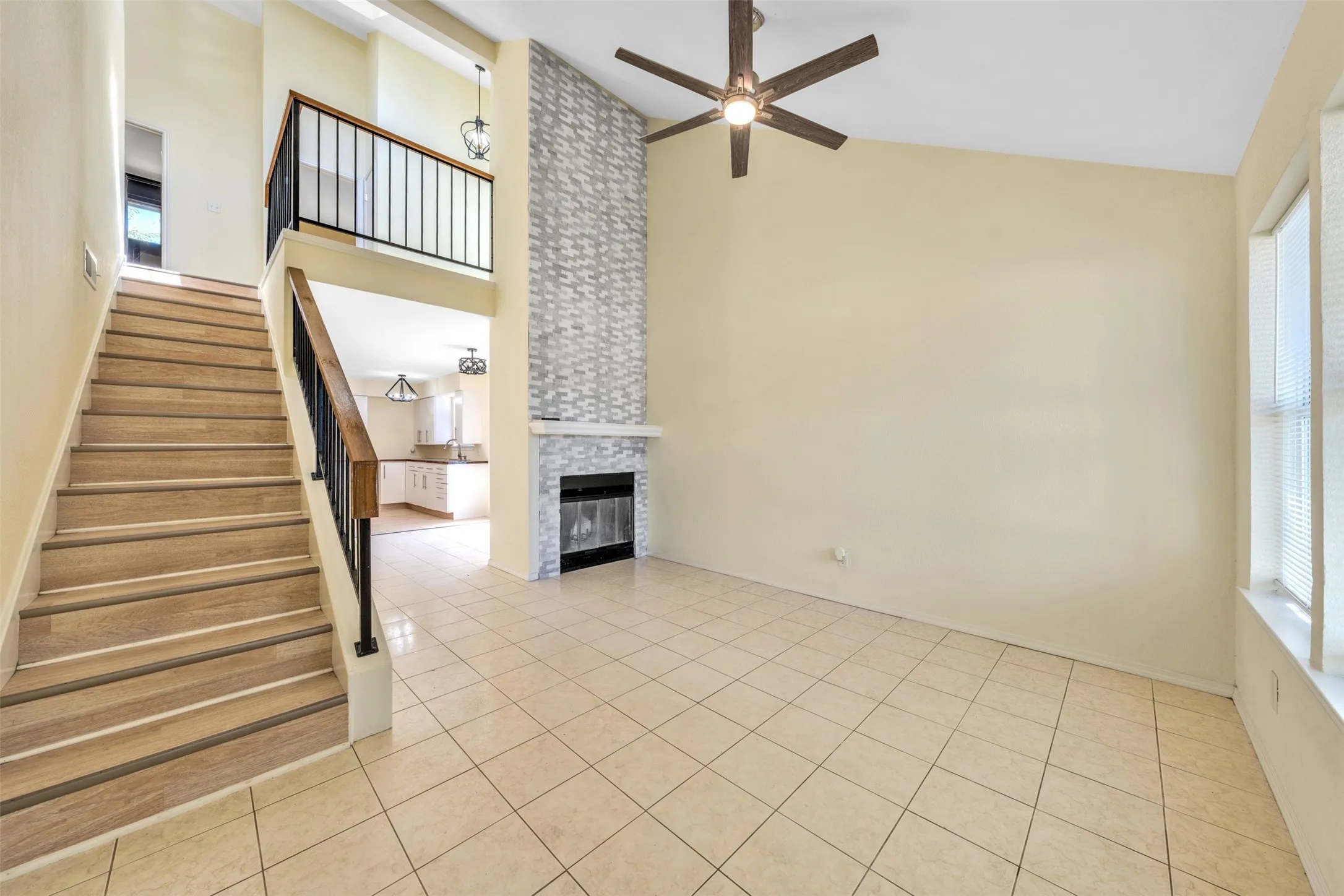 Soaring vaulted ceiling with a ceiling fan, tiled flooring, wood-finish staircase with wrought iron railing, and a fireplace with a stone-tile surround