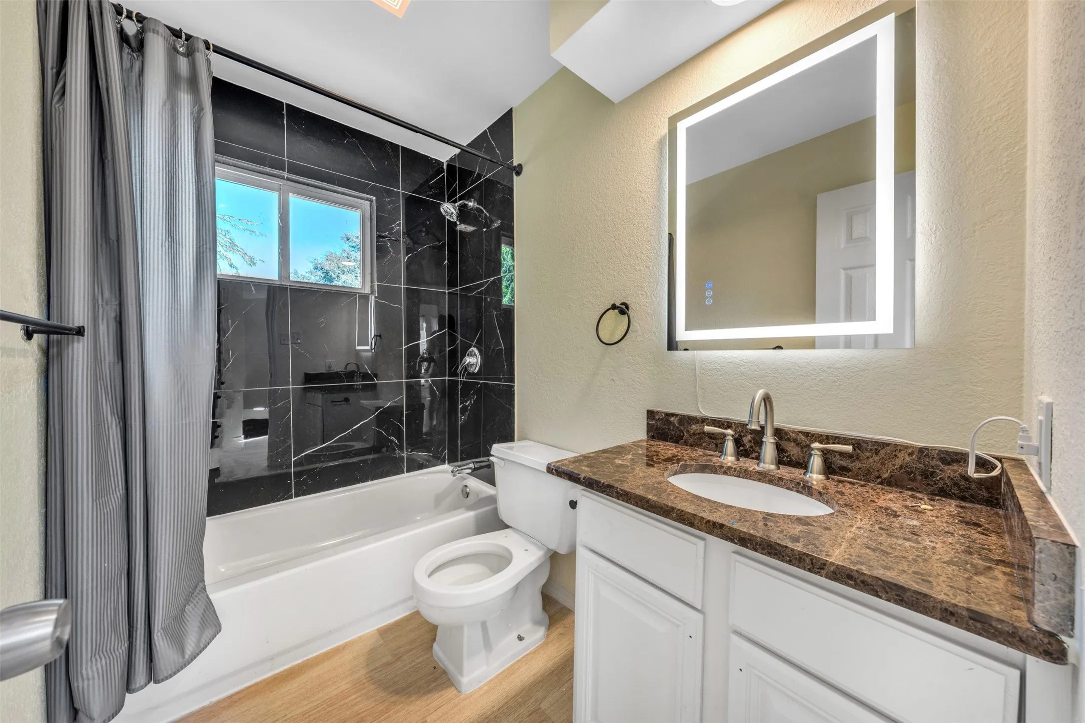 Bathroom featuring a tiled shower surround, integrated bathtub, wood-finish flooring, white vanity with a dark countertop, and an illuminated mirror