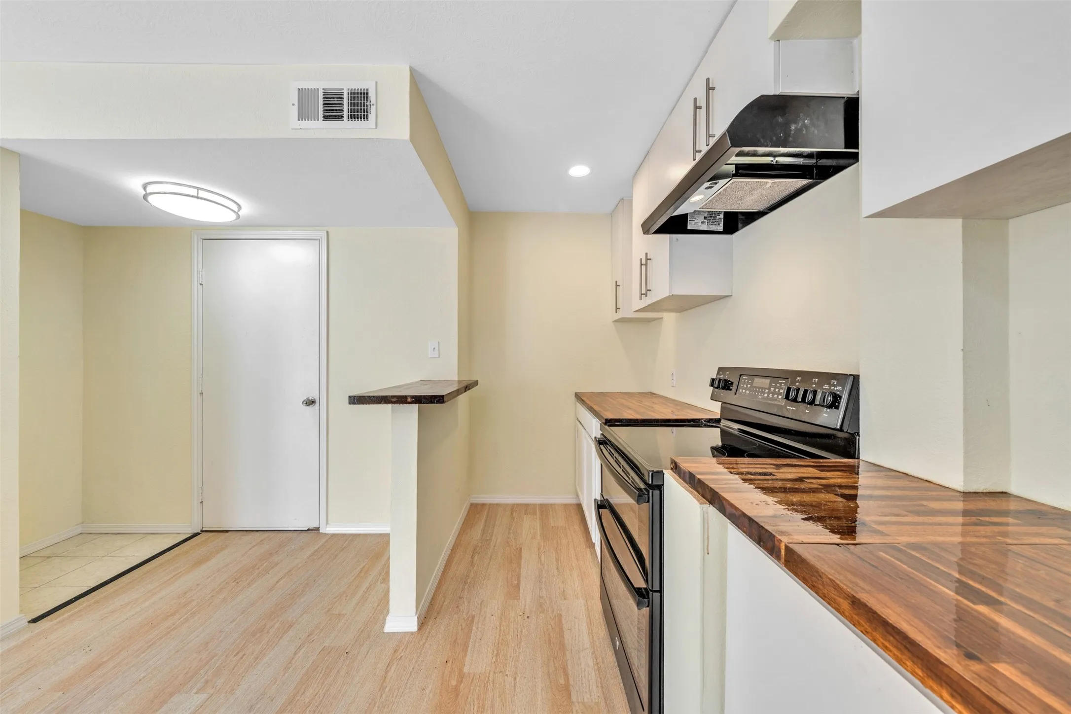 Wood-finish flooring, white cabinetry with contemporary hardware, wood-grain countertops, black range, and an overhead lighting fixture