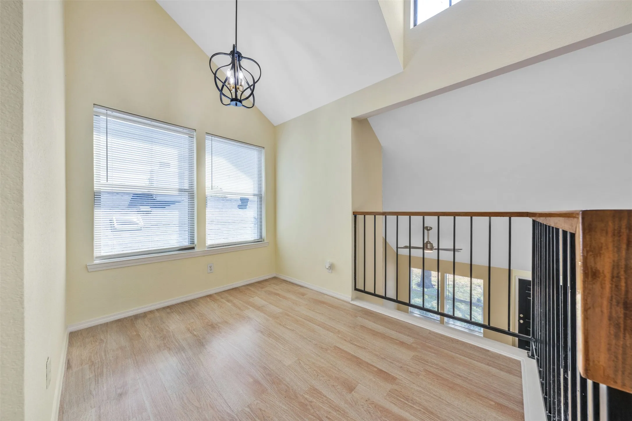 Vaulted ceiling with a decorative light fixture, two windows with blinds, wood-finish flooring, and a black metal railing