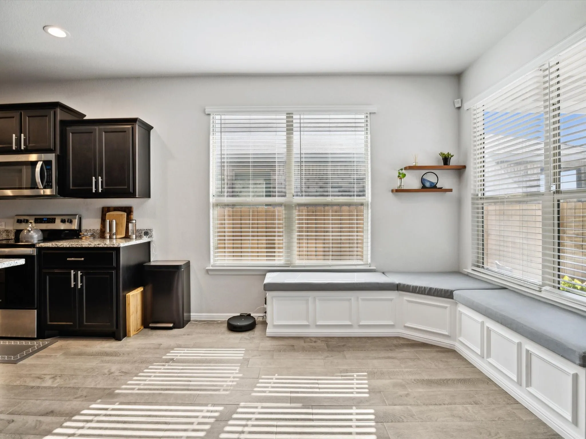 Built-in window seating with padded cushions, wood-finish flooring, dark cabinetry with stainless steel appliances, recessed lighting, and floating shelves