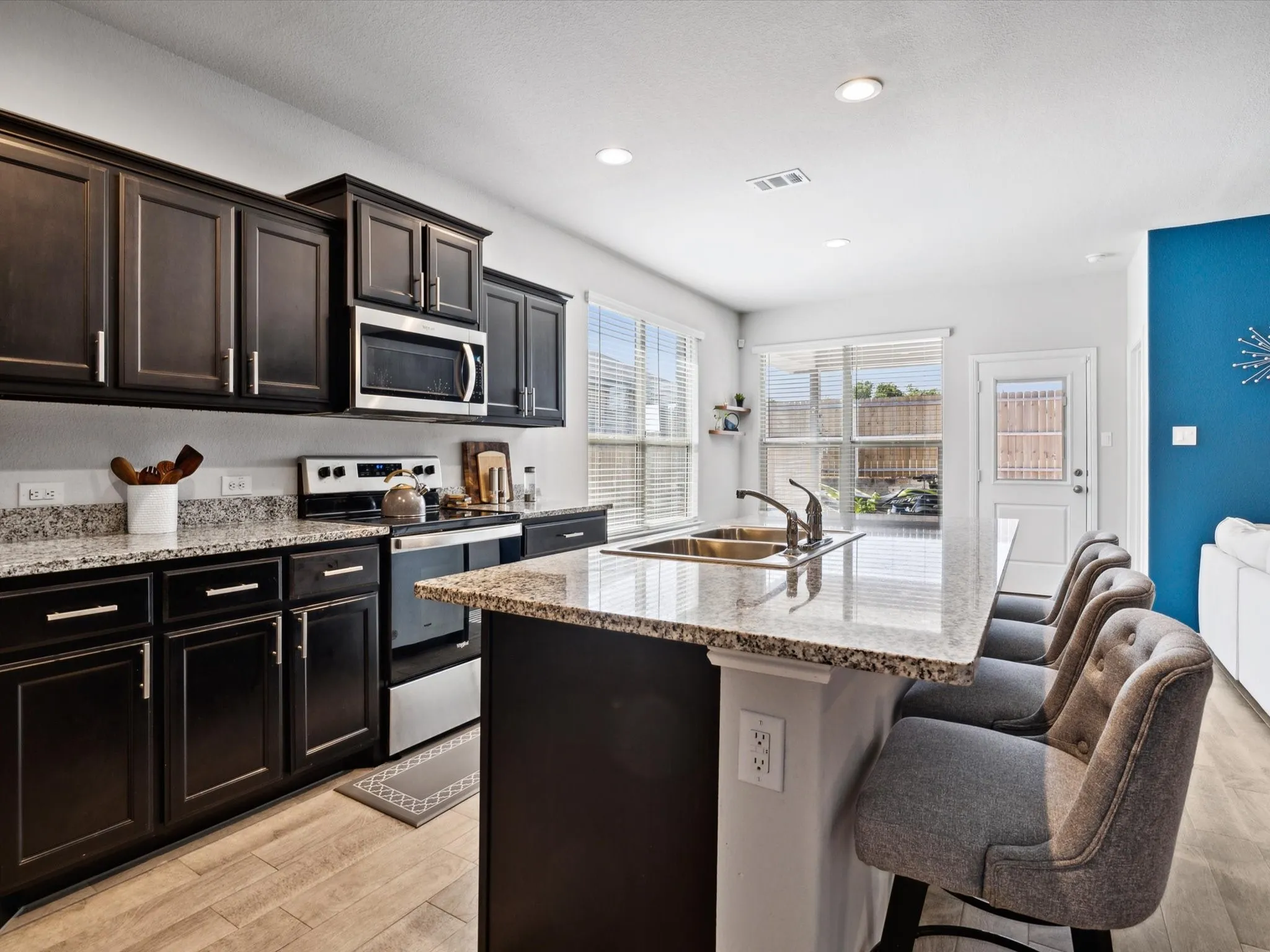 Kitchen featuring dark wood cabinetry, light-toned granite countertops, stainless steel appliances, a kitchen island with a double basin sink, and wood-finish flooring