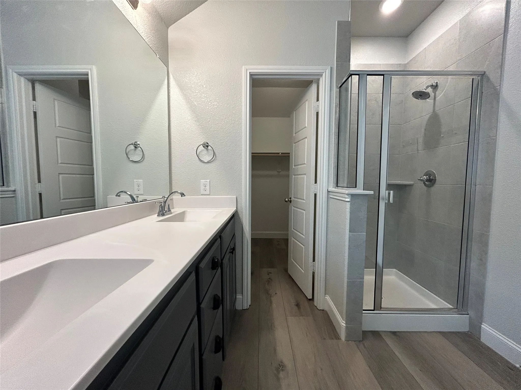 Dual vanity with integrated sinks and a continuous countertop, glass-enclosed shower with tile surround, wood-finish flooring, and a walk-in closet