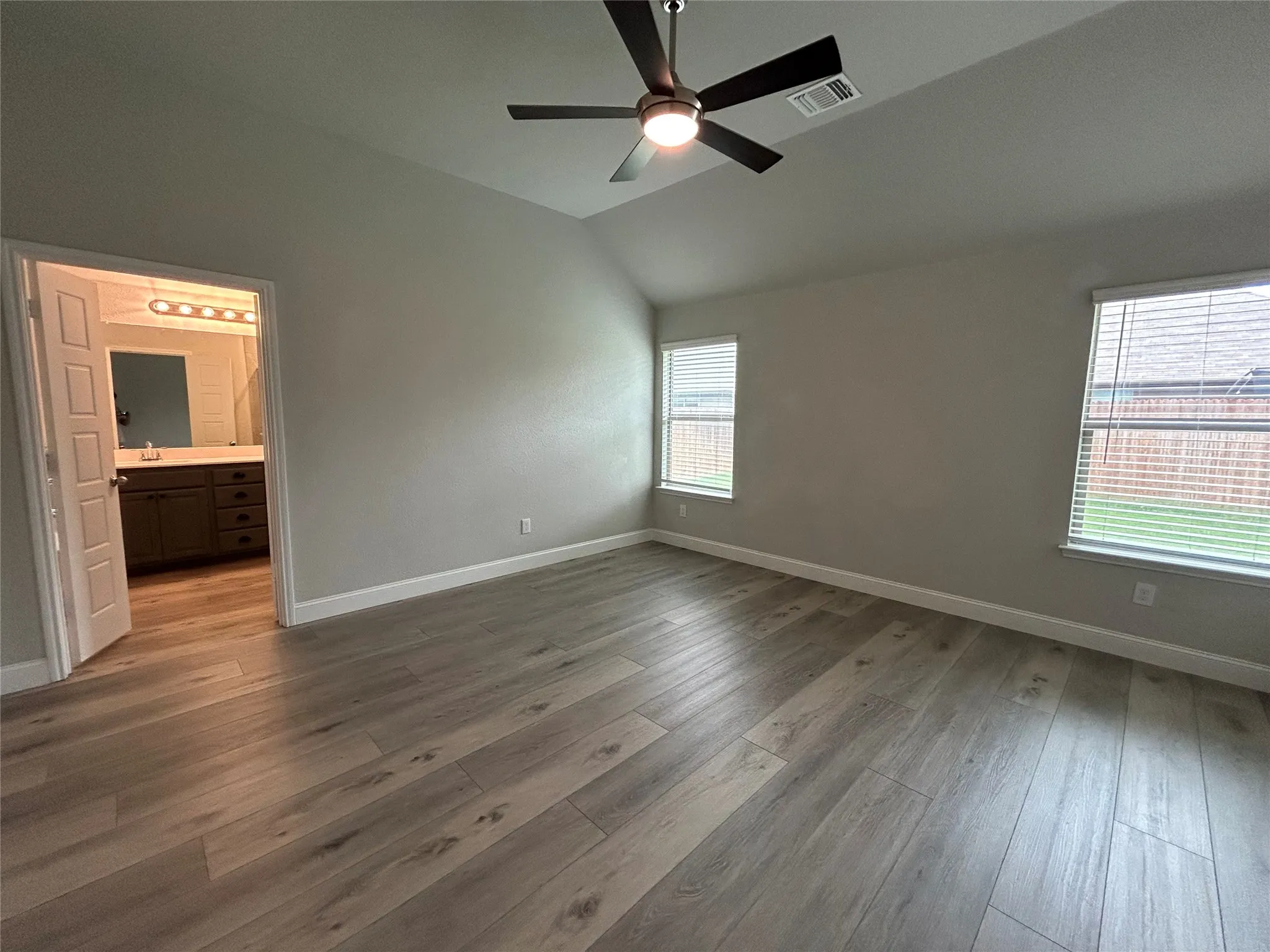 Vaulted ceiling, wood-finish flooring, two windows with blinds, ceiling fan with light fixture, and an en-suite bathroom with a vanity and mirror