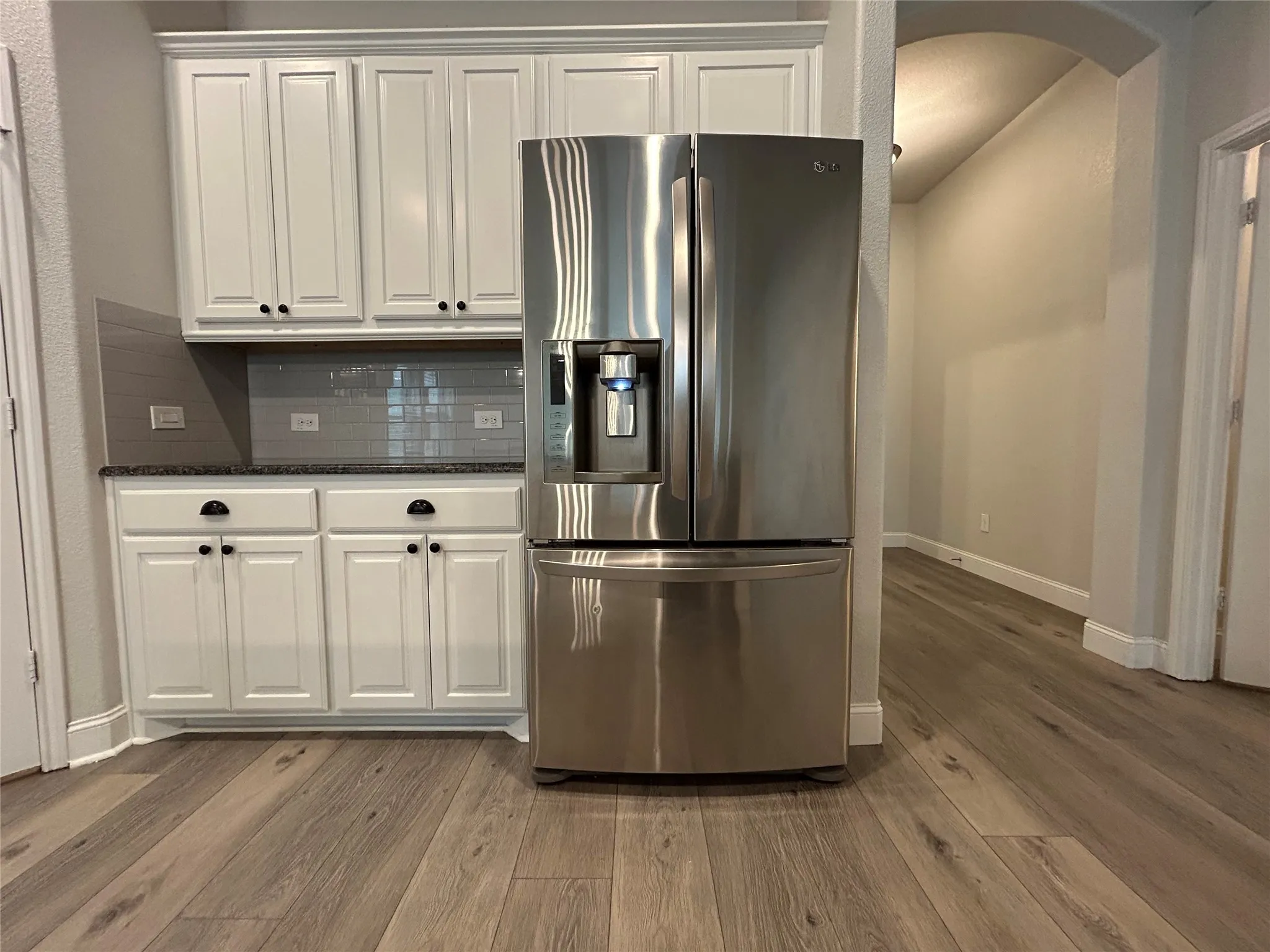White shaker-style cabinetry with black hardware, dark stone-finish countertops, subway tile backsplash, stainless steel refrigerator, and wood-finish flooring