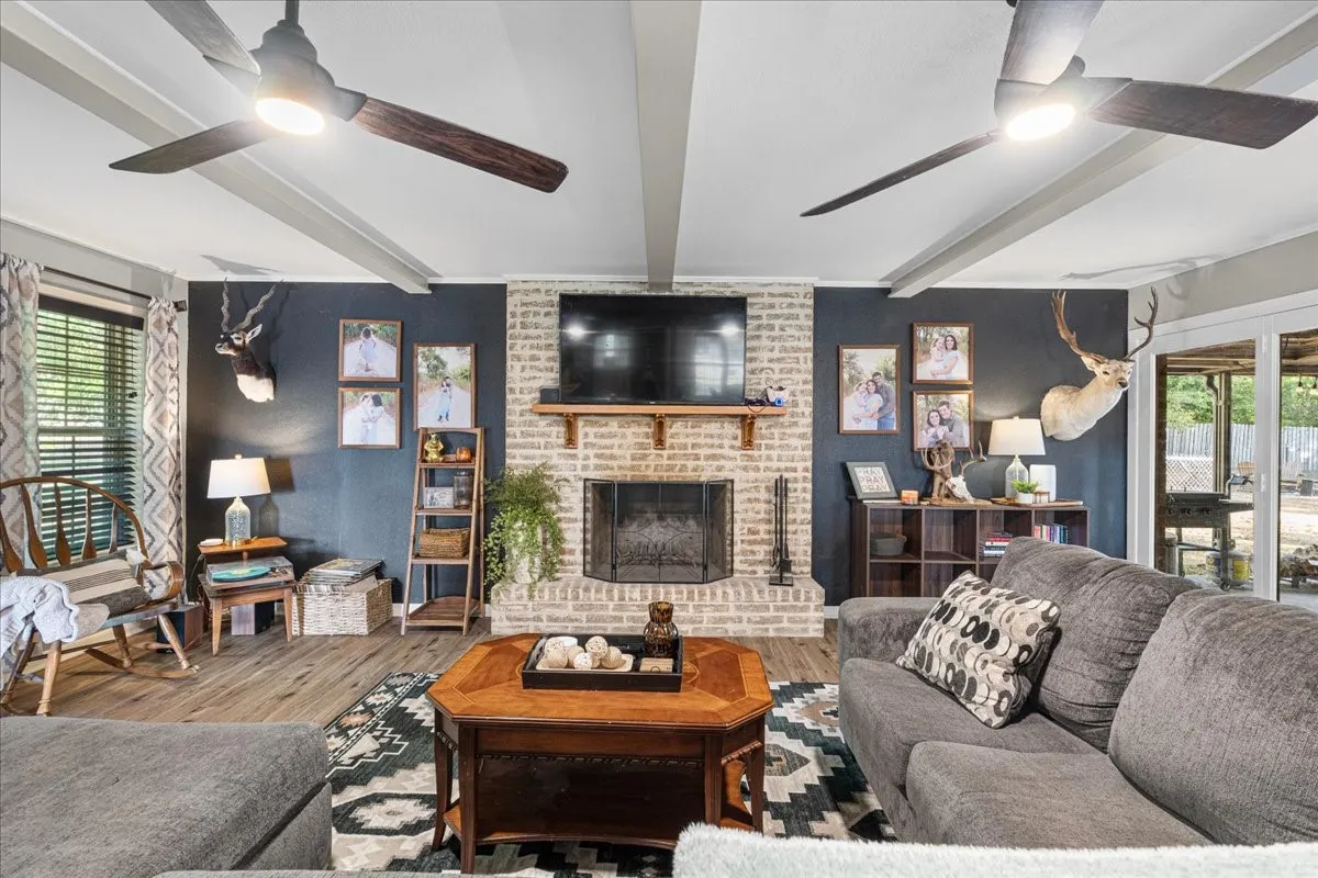 Exposed ceiling beams, brick fireplace with a wood mantle, wood-finish flooring, and dual ceiling fans