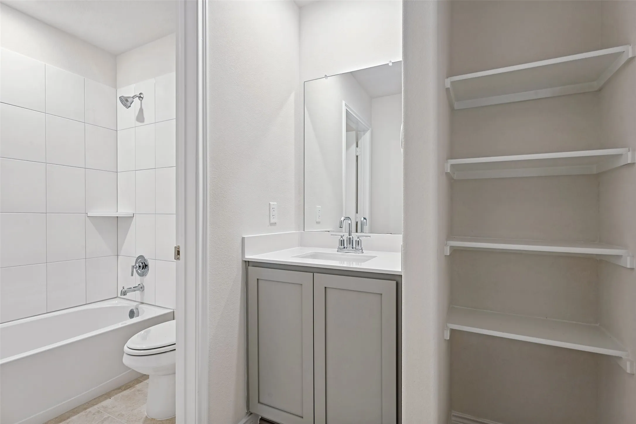 Bathroom featuring a tub/shower combination with white tile surround, a vanity with a quartz-look countertop, a rectangular mirror, and built-in shelving
