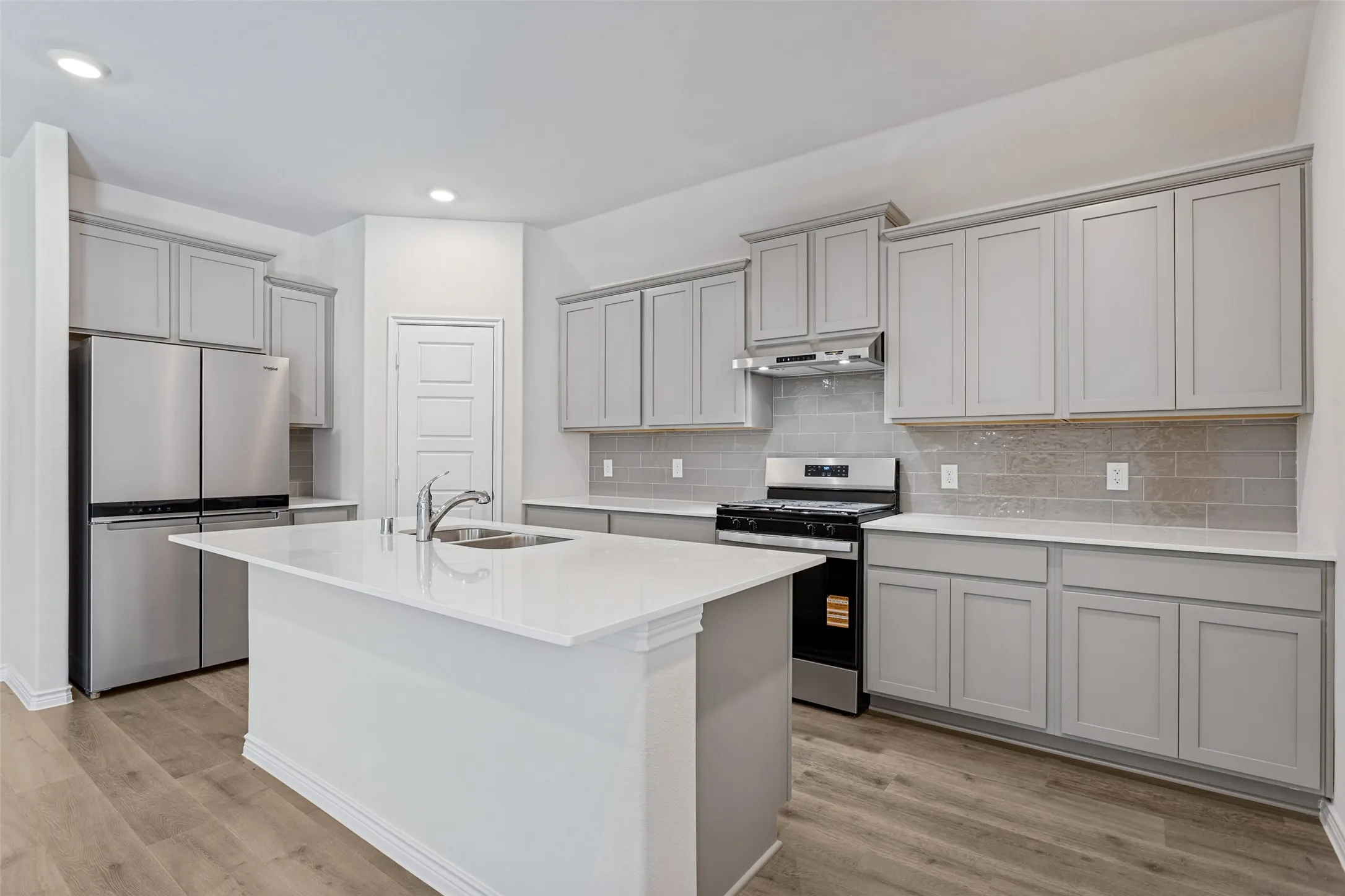 Kitchen featuring an island with an undermount sink, stainless steel appliances, light gray cabinetry, white countertops, and wood-finish flooring