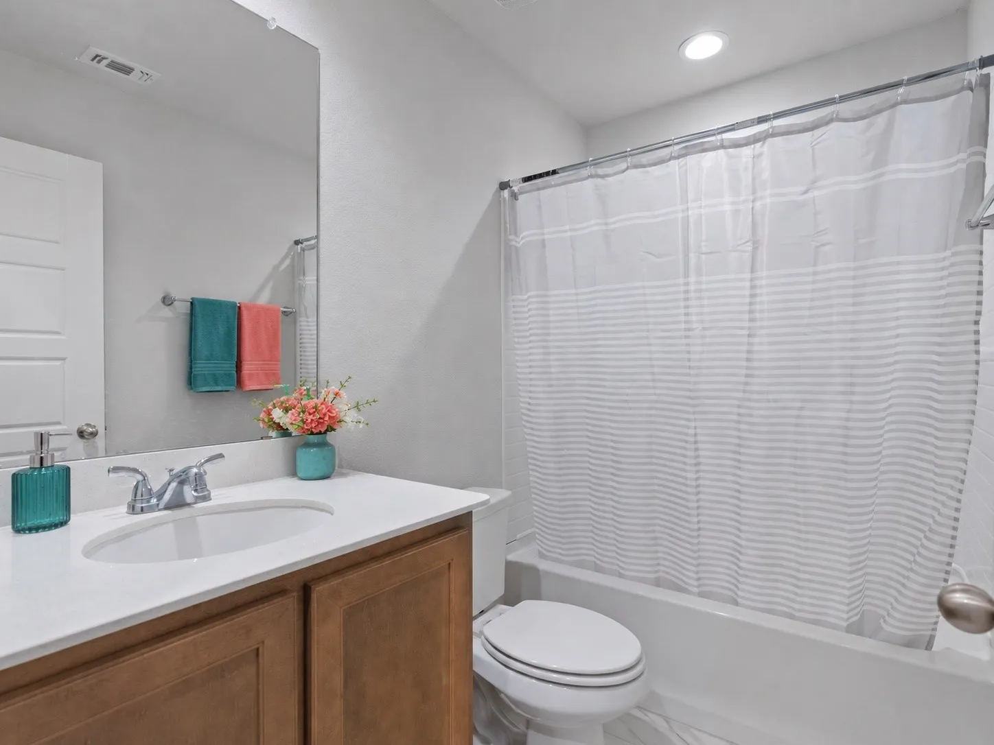 White wall-mounted mirror, wood-finish vanity with a white countertop, chrome faucet, white bathtub with a shower rod, and recessed ceiling lighting